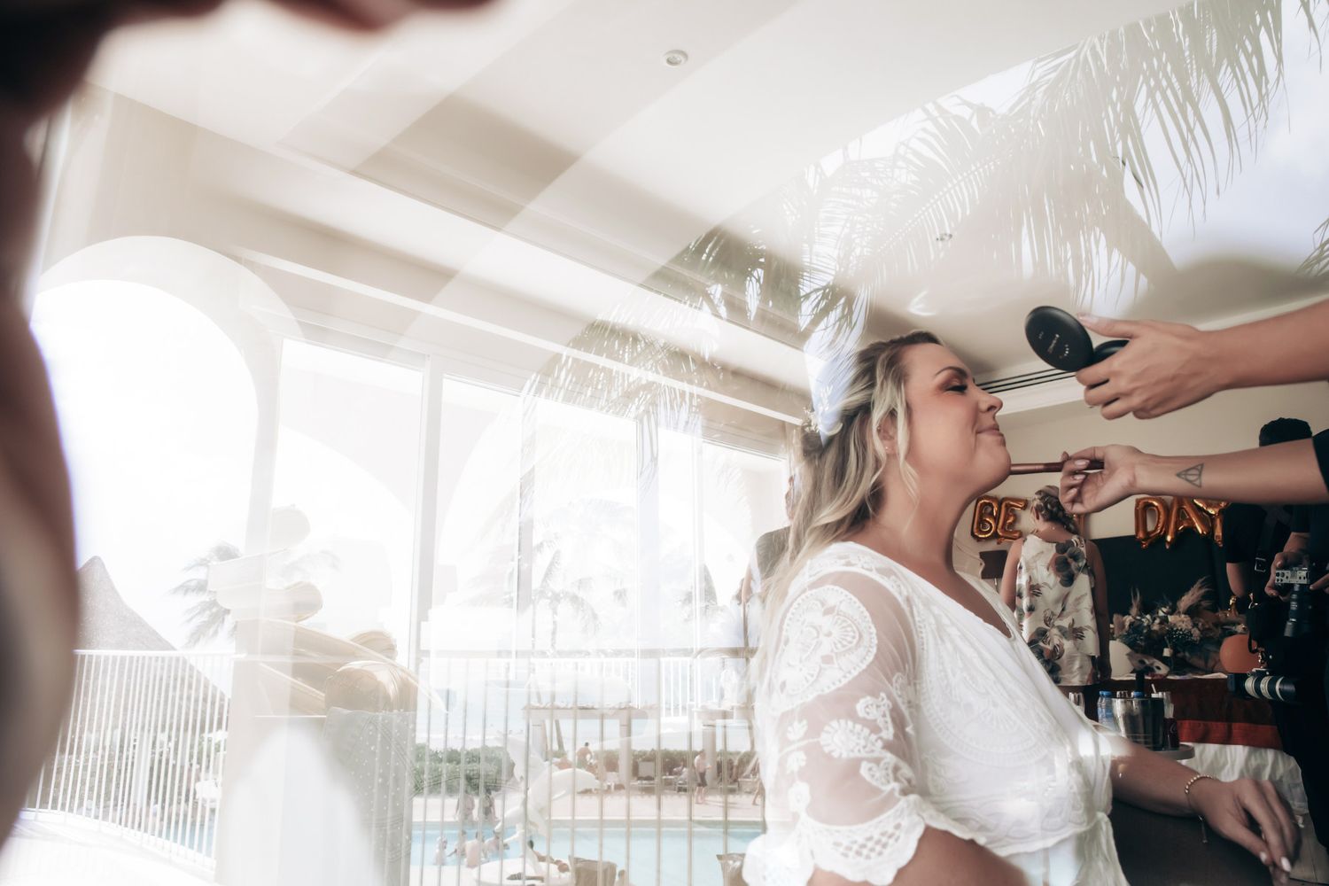 Bride in white lace dress getting hair styled indoors with natural light and pool visible in background.