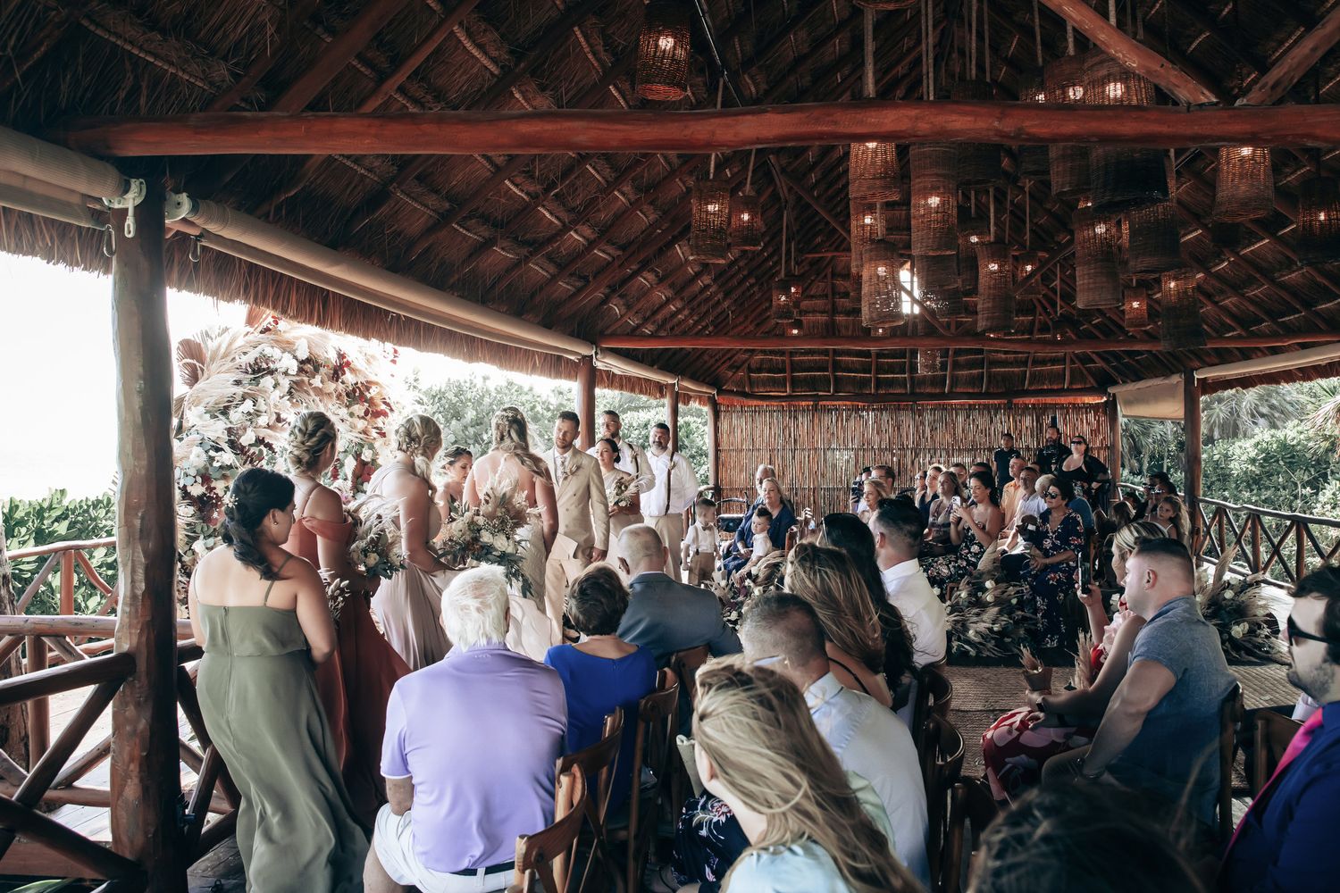 Guests gather at rustic open-air pavilion wedding ceremony surrounded by lush tropical greenery.