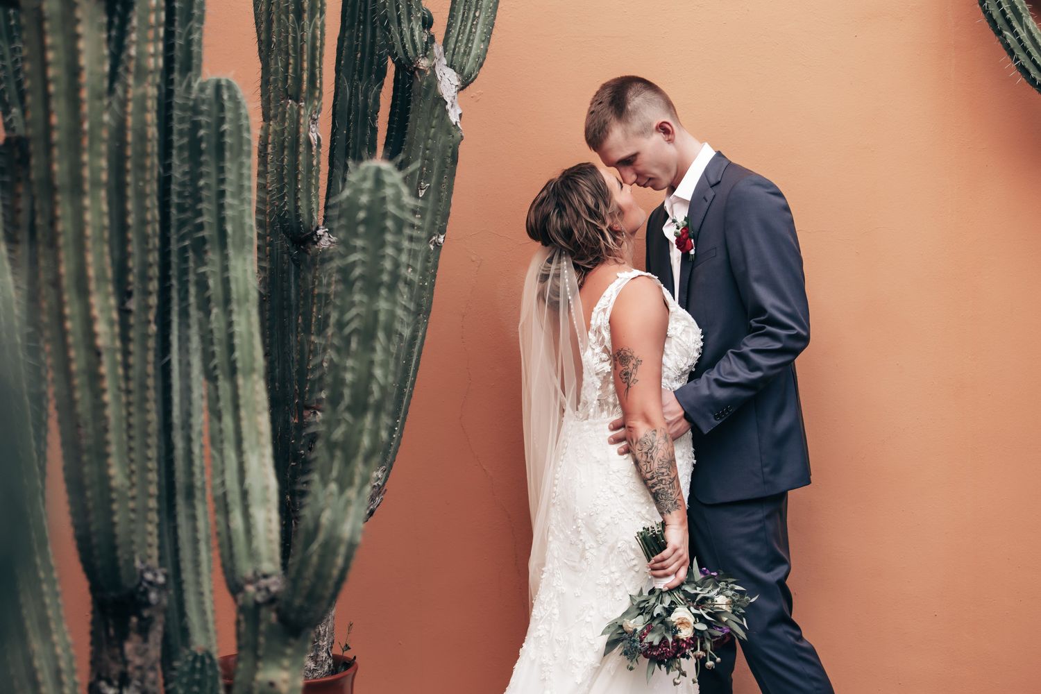 Bride and groom share an intimate moment beside tall cacti against a warm terracotta wall.