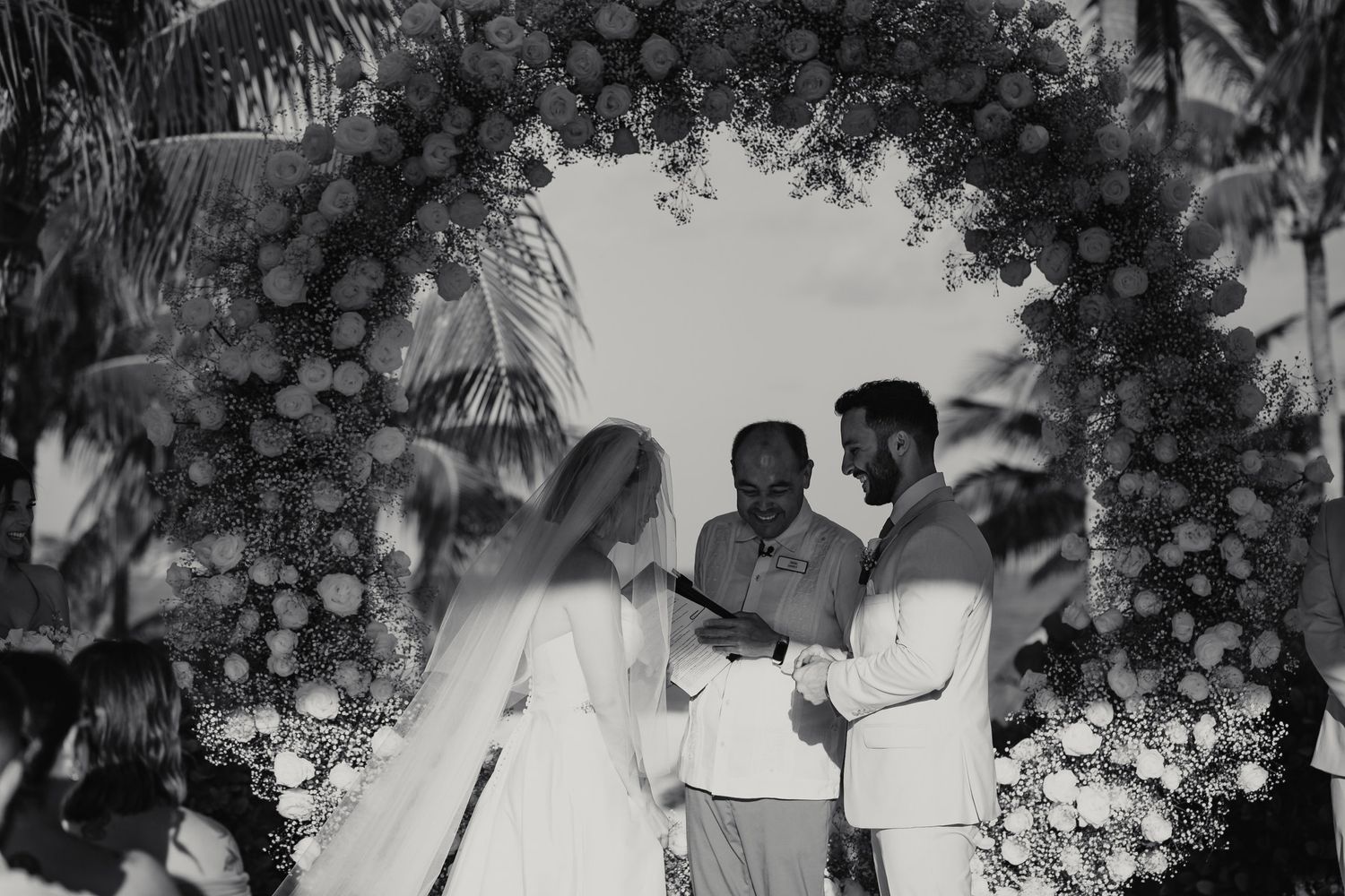 A couple exchanges vows under a floral arch at an outdoor wedding ceremony in black and white.