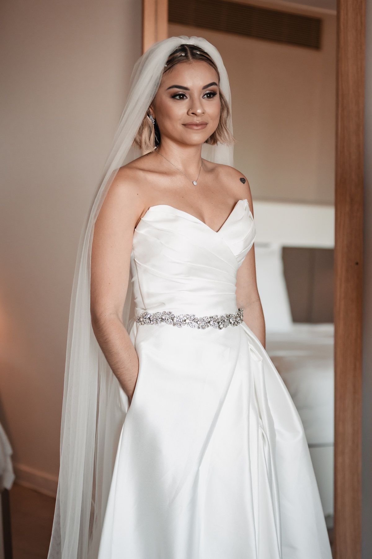 Elegant bride in strapless satin wedding gown with crystal belt and long veil posing indoors.