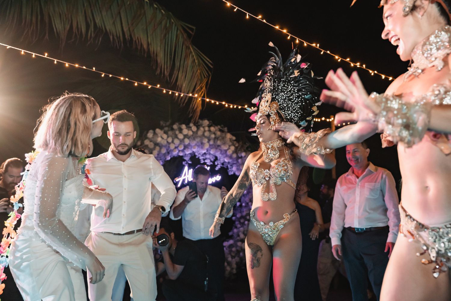 Bride and groom dance with samba performers in feathered costumes at tropical wedding reception.