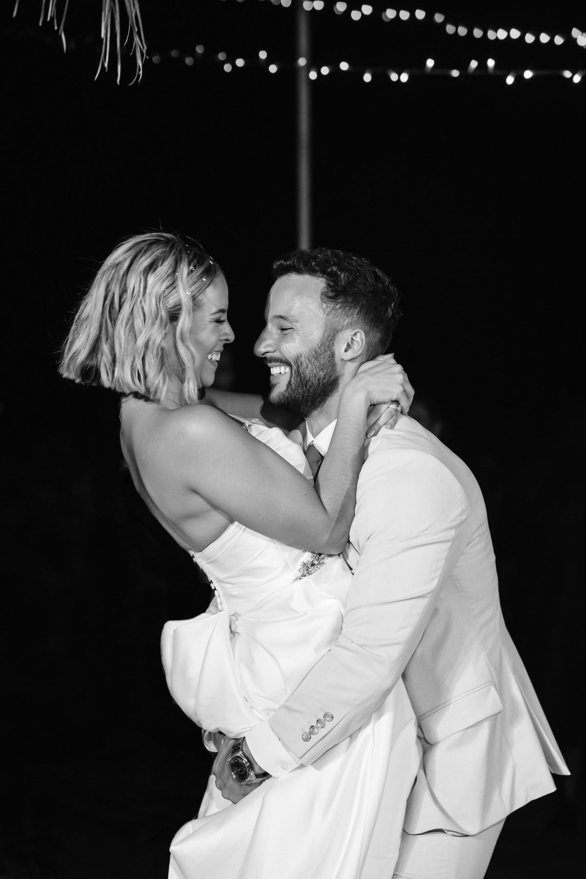 Black and white photo of groom lifting laughing bride during joyful wedding reception dance at night.