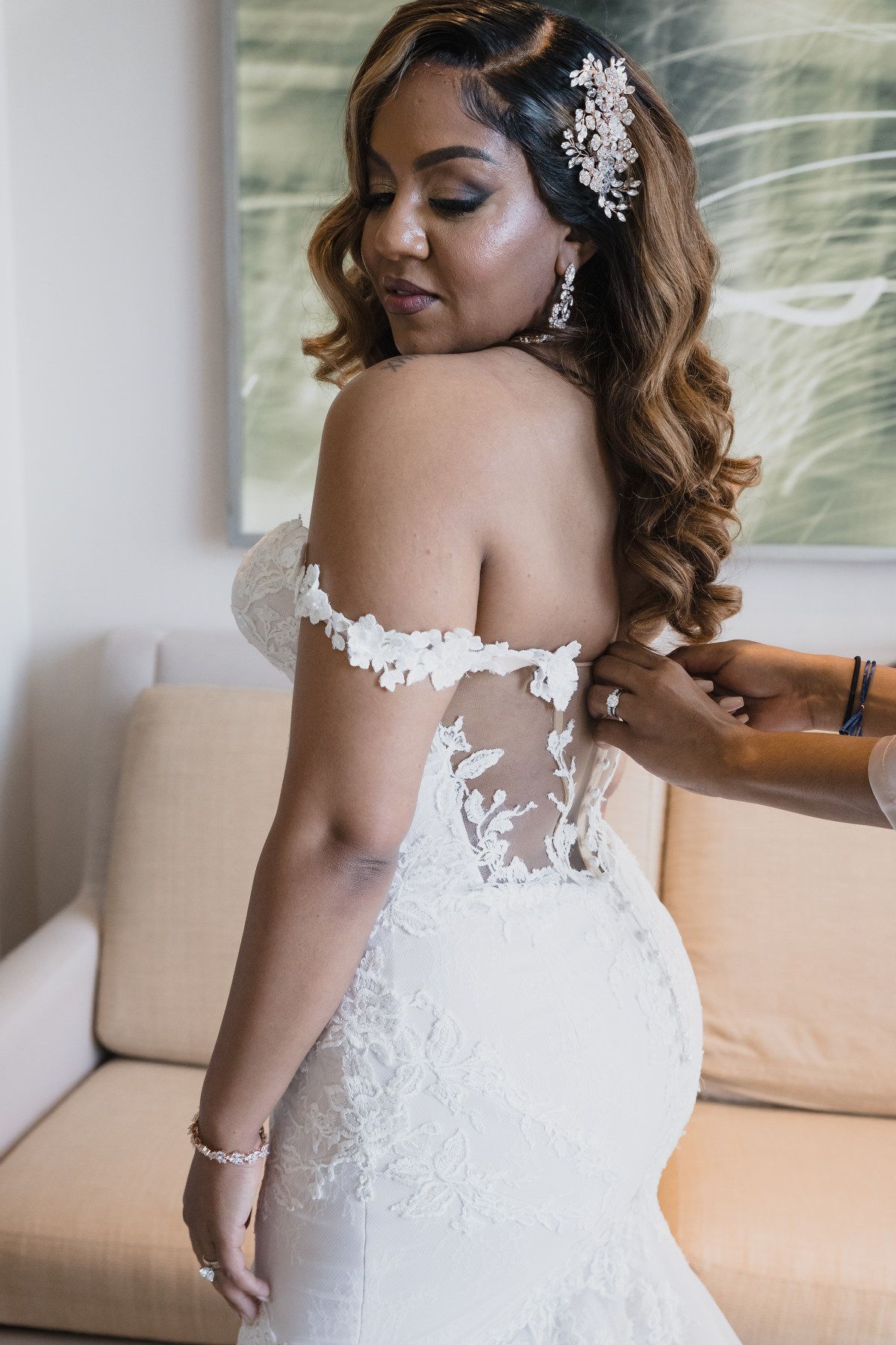 Bride in off-shoulder lace mermaid gown with floral appliques being buttoned up by a bridesmaid.