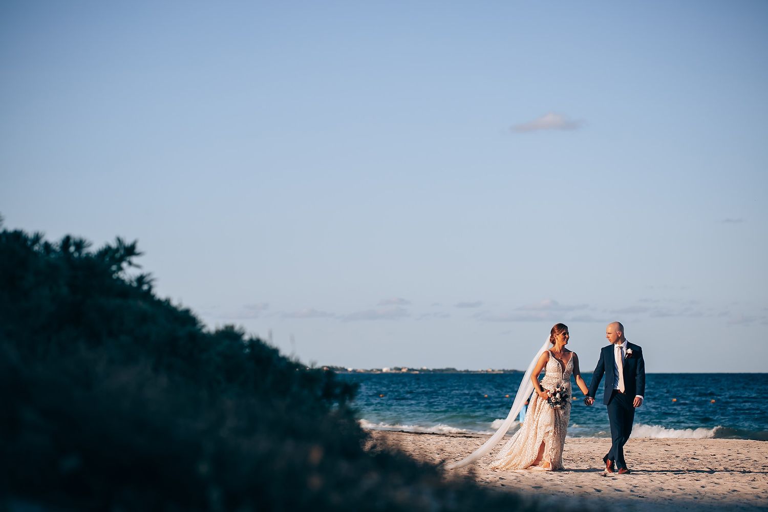 A bride and groom walk together on a beach with palm trees in the background.