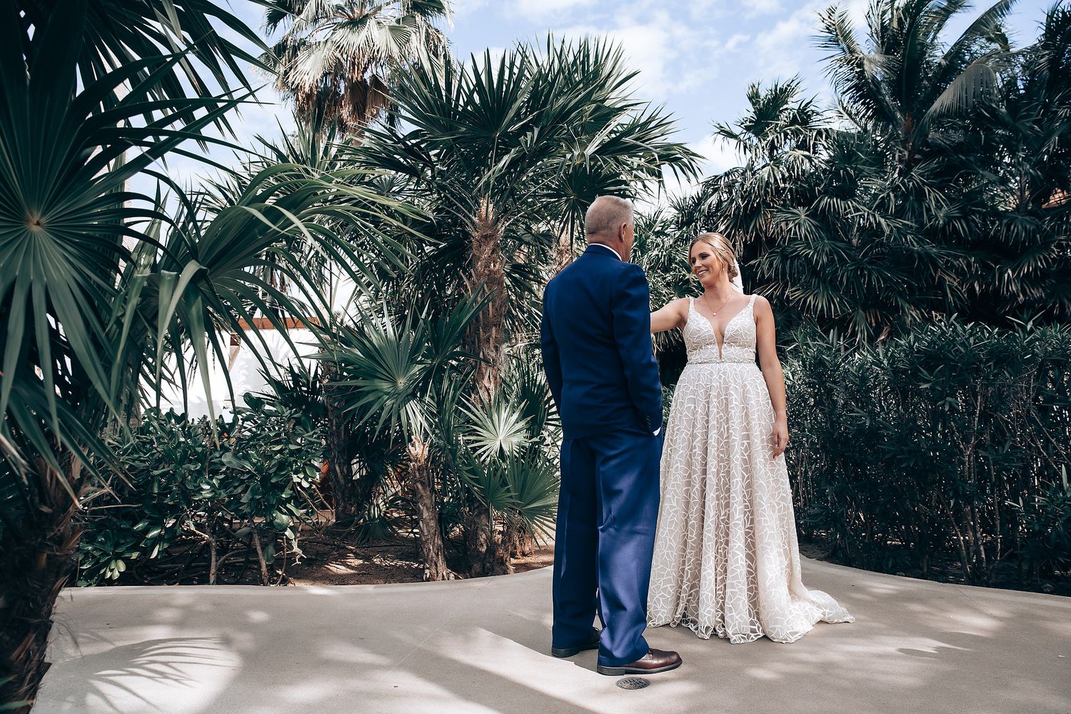 A bride and groom stand together in a tropical garden setting, surrounded by palm trees.