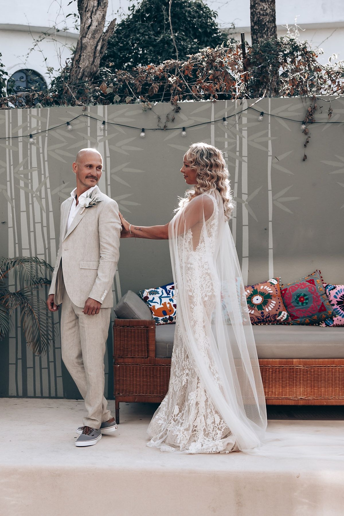 A couple stands together in a room with a wicker bench and decorative wall art.