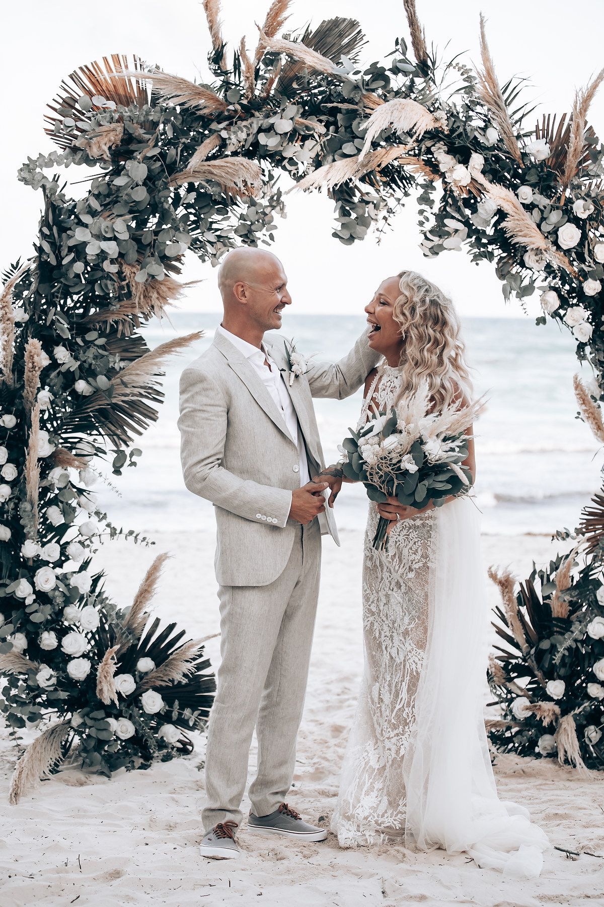 Elegant beach wedding scene with floral arch decorations and pampas grass against an ocean backdrop.