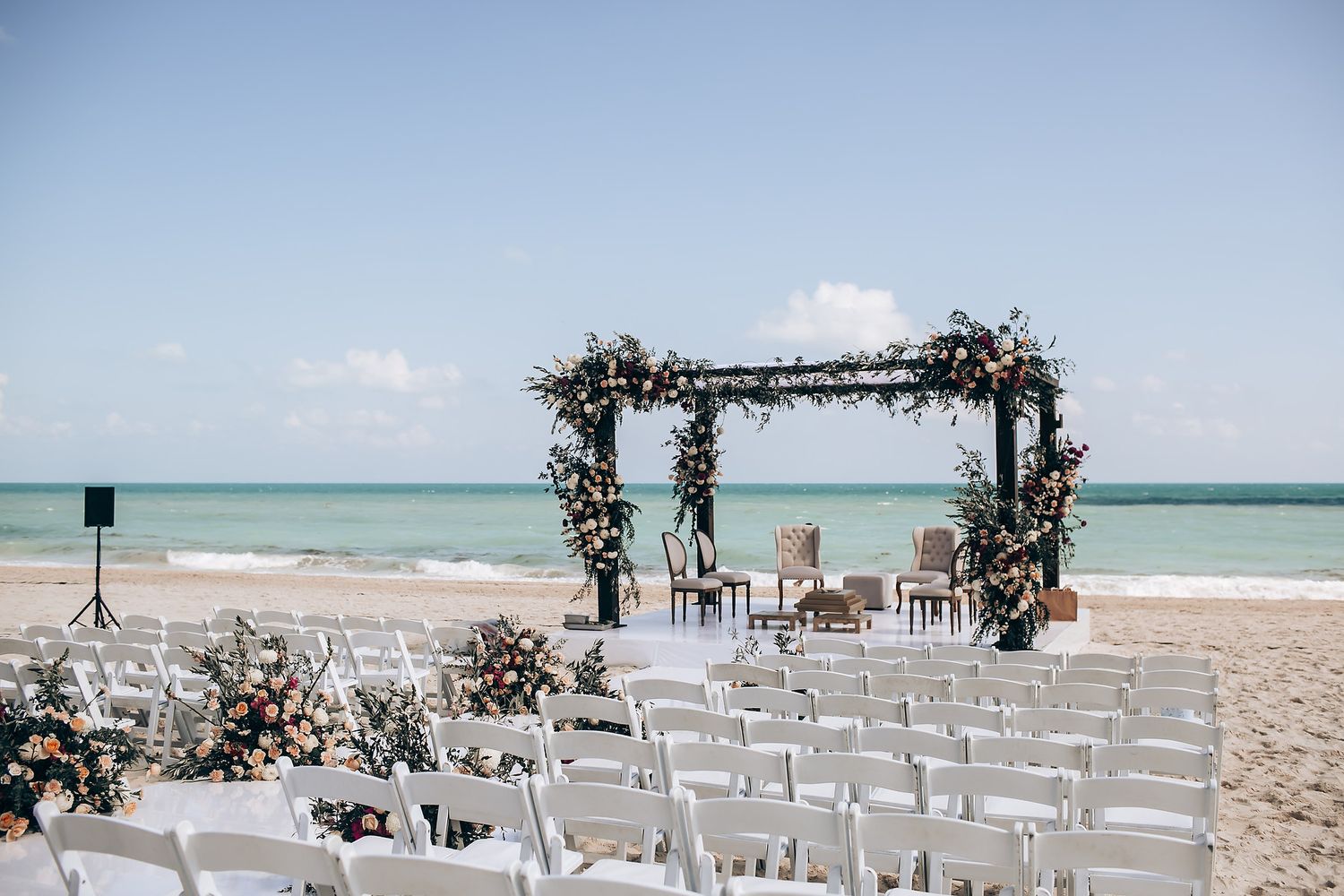 Beach wedding setup with floral arch and white chairs overlooking the ocean.