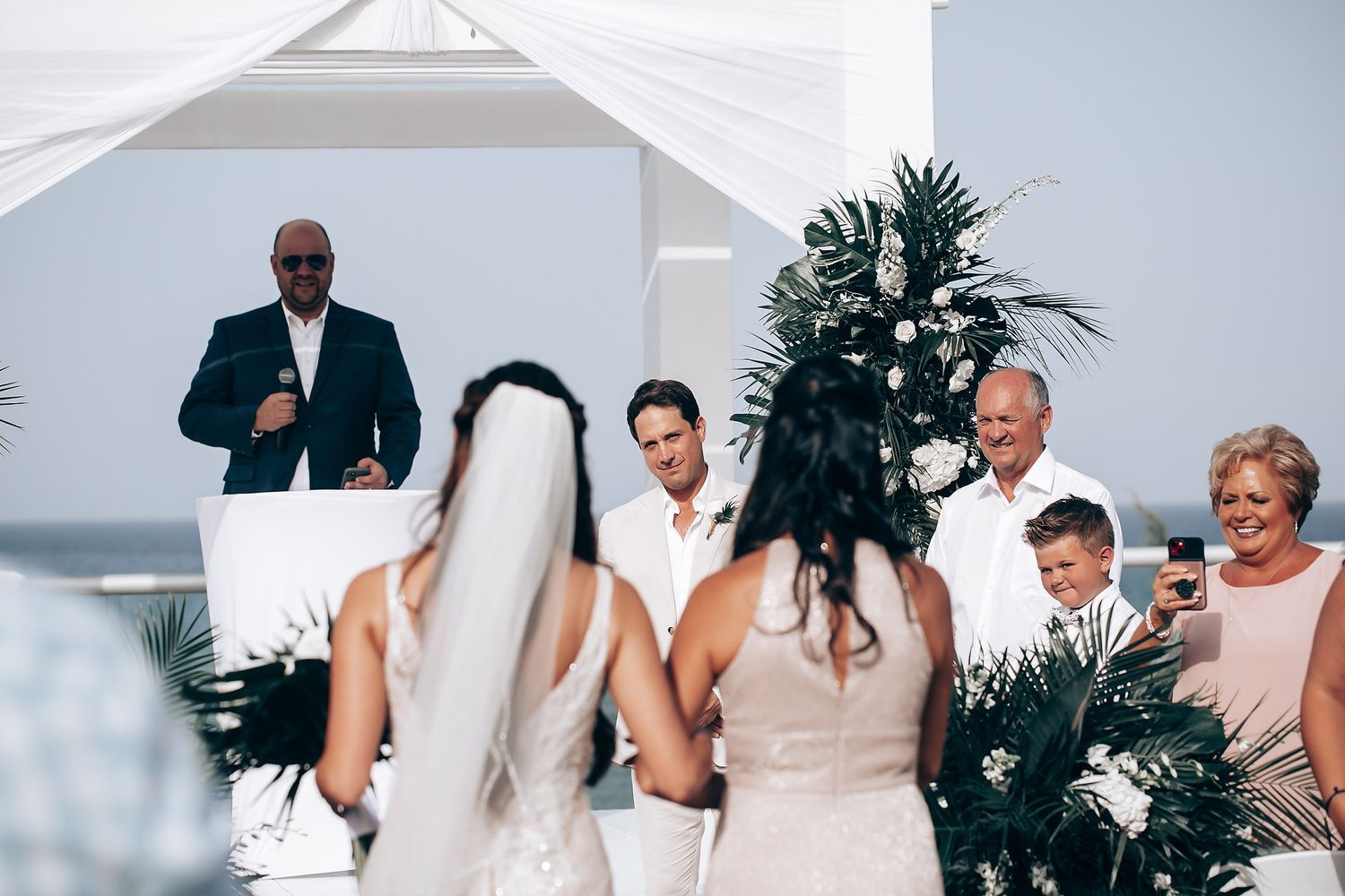 Wedding guests seated in white chairs on a beachfront terrace with tropical palm decorations and ocean views.