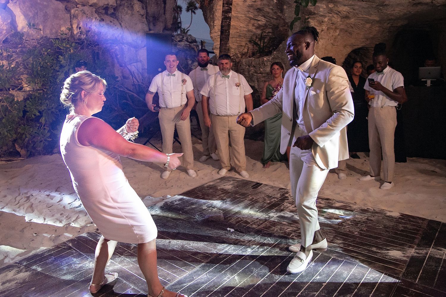 Wedding party dancing on illuminated floor in cave-like venue with purple lighting.