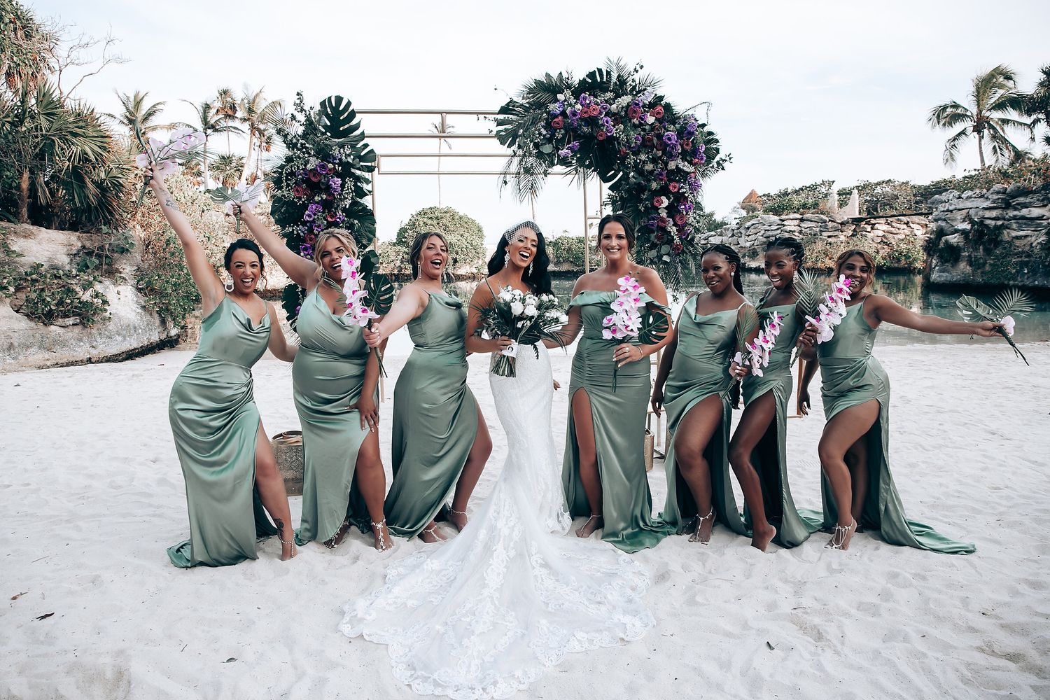 Wedding party celebrates joyfully on beach with bridesmaids in matching sage dresses.