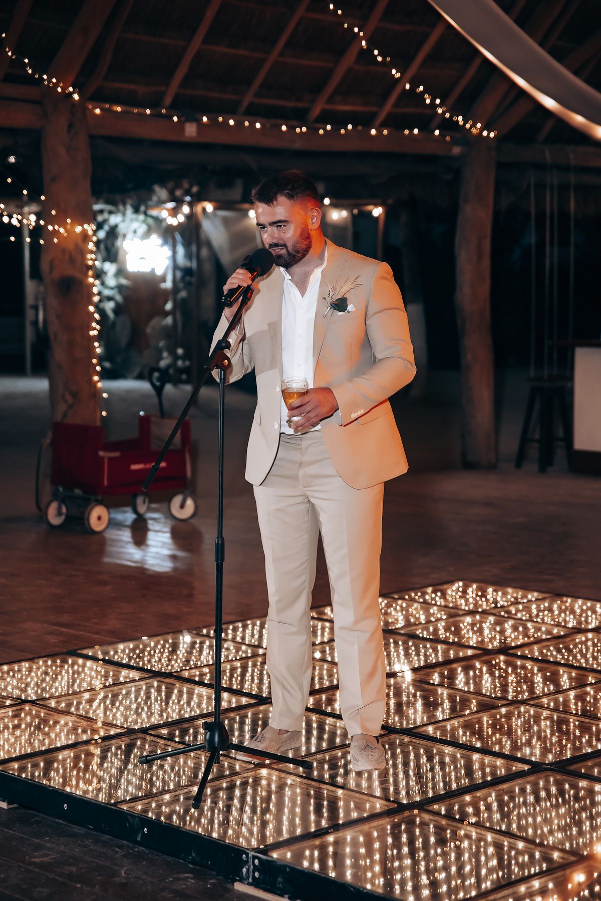 A person in a light colored suit speaks into a microphone surrounded by glowing string lights in a rustic venue setting.