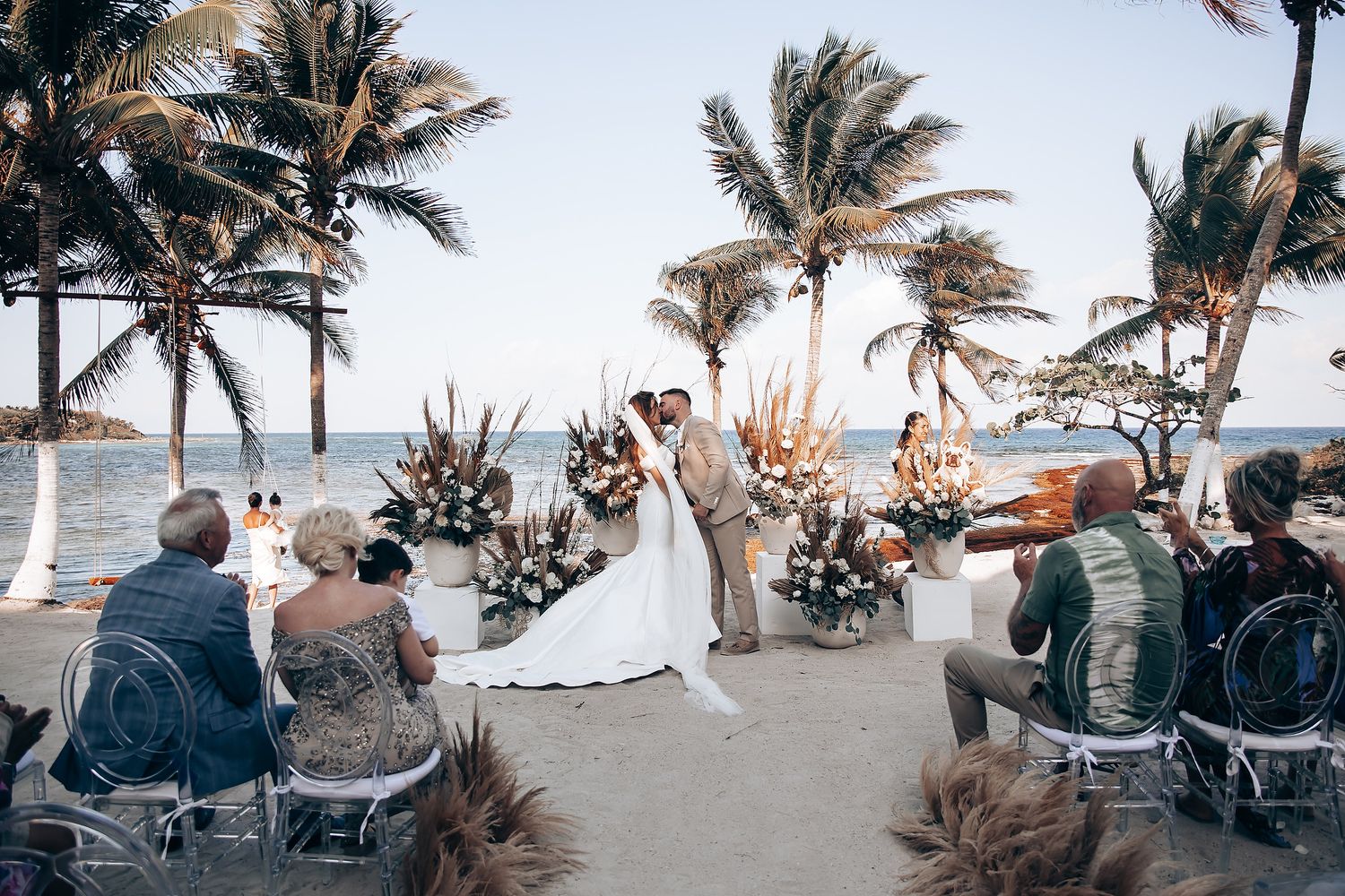 Wedding guests seated in white chairs on a tropical beach with palm trees swaying in the wind during sunset ceremony.