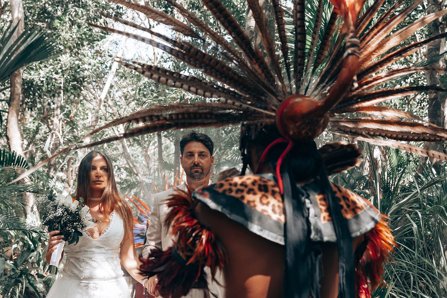 Traditional Aztec dancer performs in ceremonial feathered headdress and regalia alongside couple in outdoor setting.