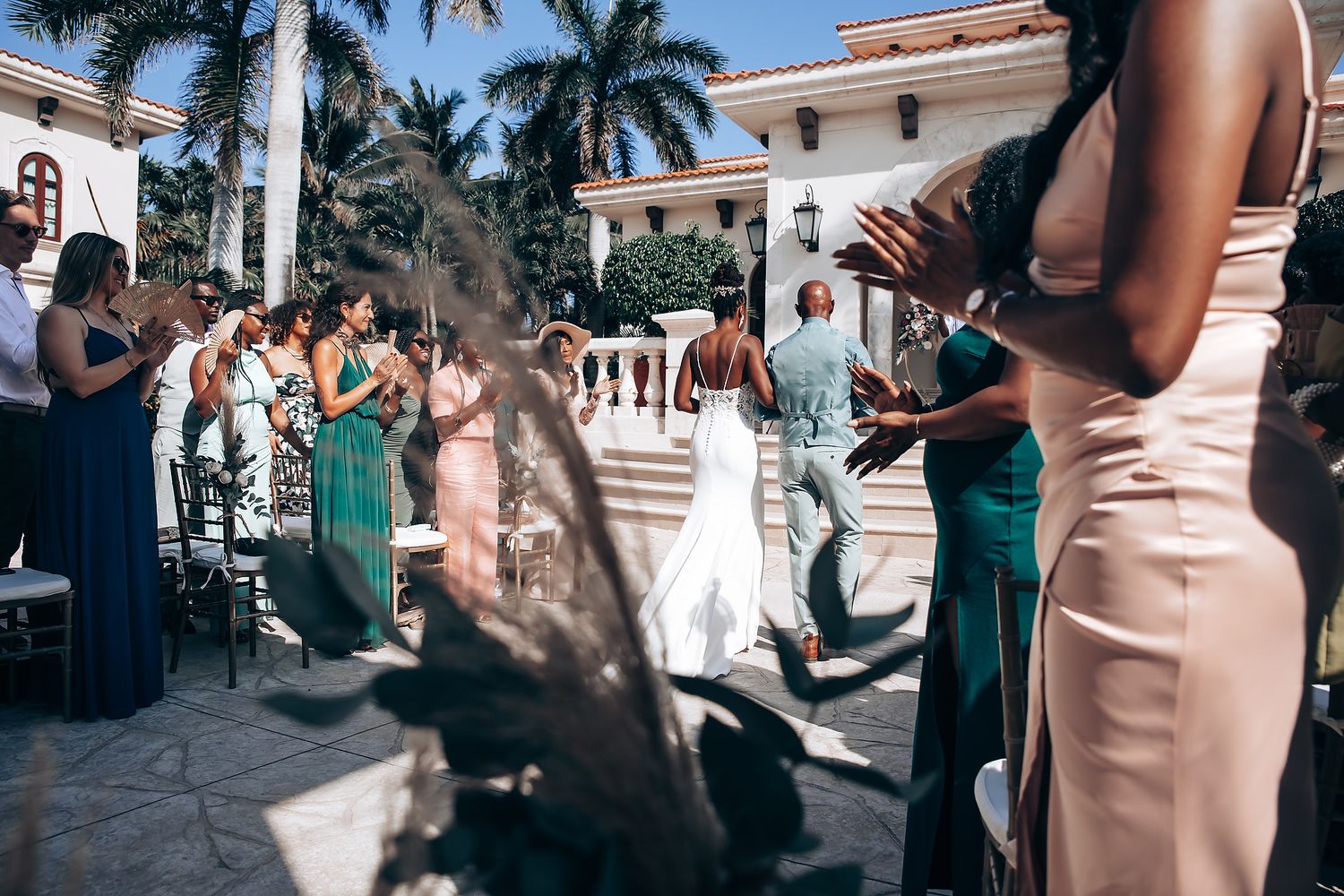 Wedding party in teal and blush dresses poses together at tropical venue with palm trees.
