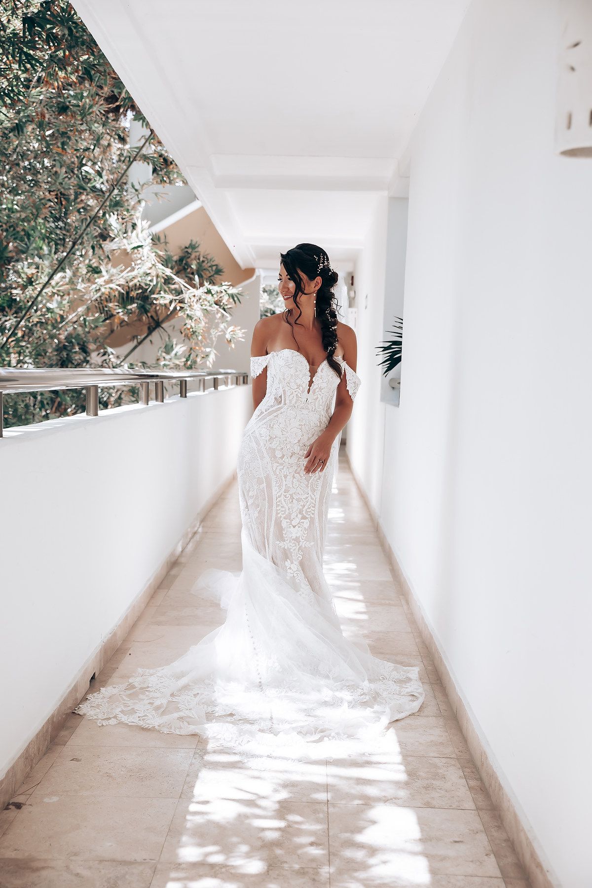 A bride in a white lace wedding dress walks down a bright sunlit hallway with dappled shadows on the floor.