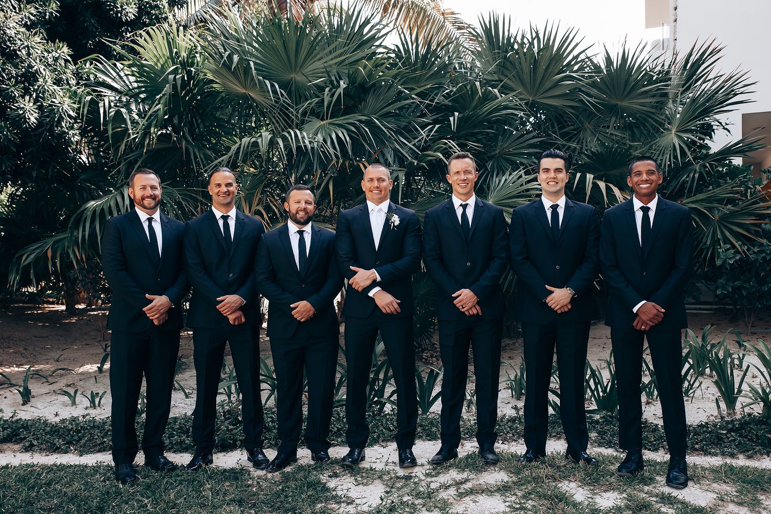 Groomsmen in black suits pose together outdoors with palm trees in the background.