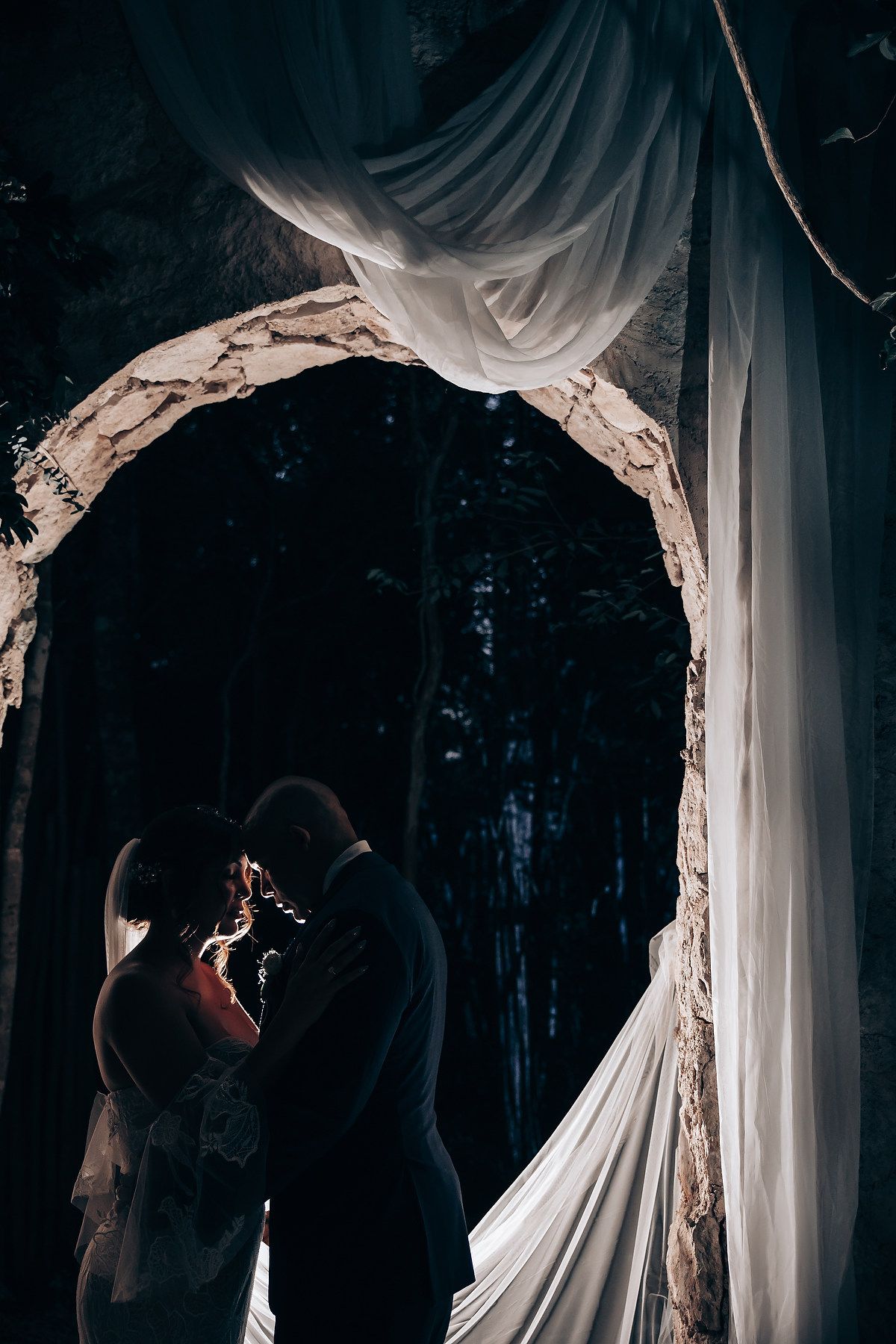 Romantic silhouette under draped white fabric arch in forest setting.