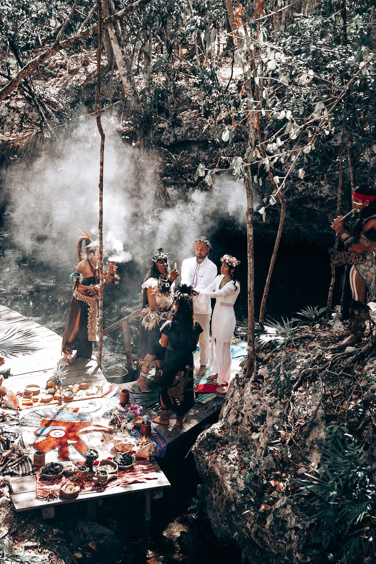 Traditional ceremony takes place in a forested setting with participants and ritual objects laid out on ground.