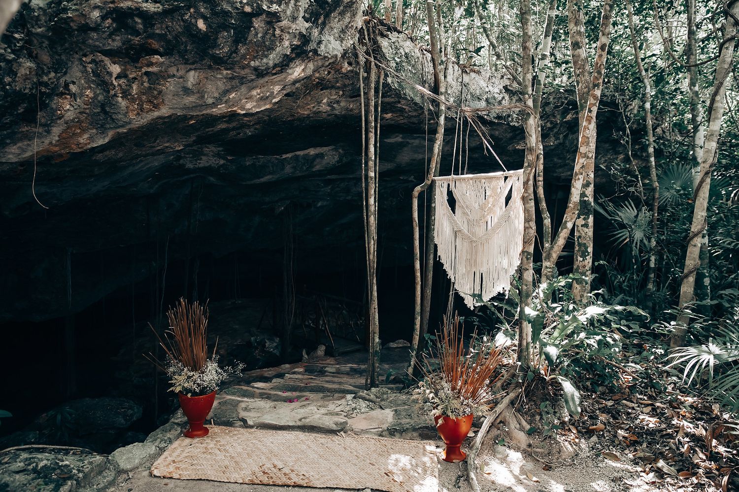 A white hammock hangs in front of a dark cave entrance with red flowers decorating the ground.