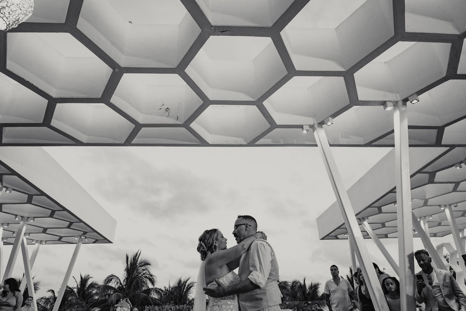 Black and white photo of bride and groom sharing first dance under geometric honeycomb ceiling.