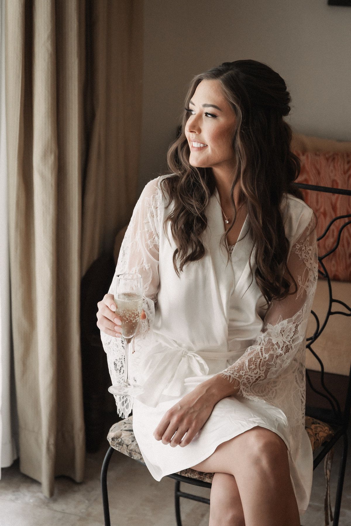 Beautiful bride in a white lace robe holding a champagne glass while getting ready for her wedding.