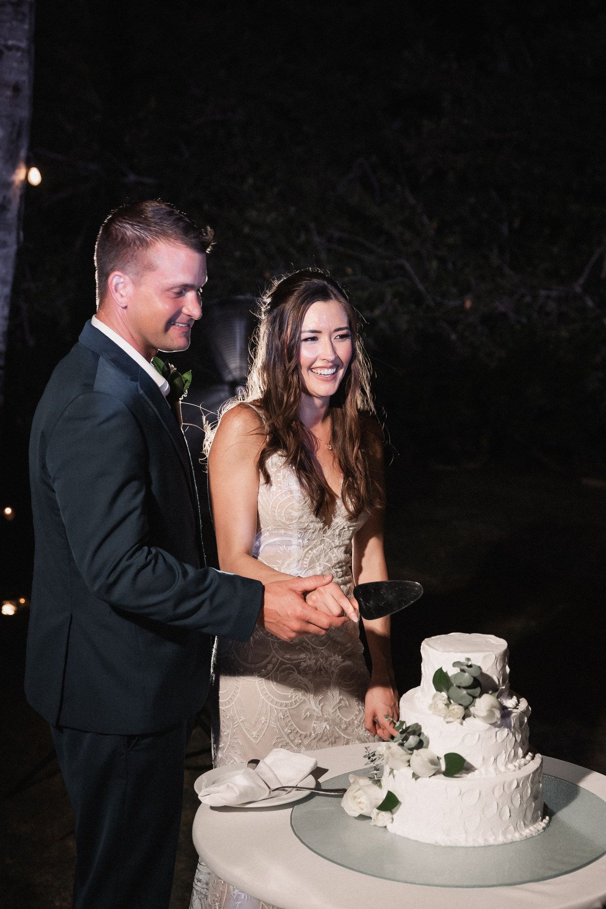 Smiling bride and groom cutting white two-tier wedding cake decorated with greenery at night reception.