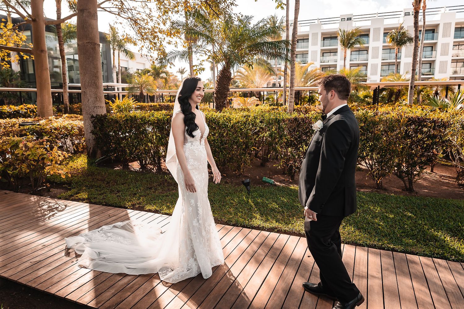 Bride in white gown and groom in black suit share a first look on a wooden deck surrounded by lush greenery.