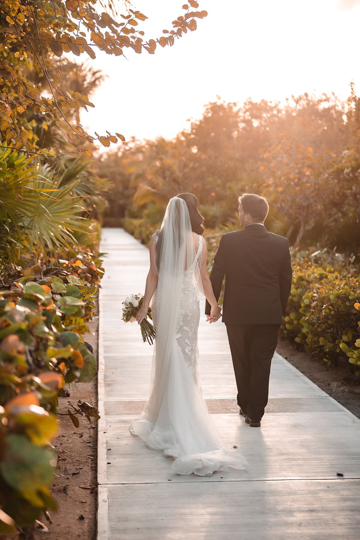 Bride and groom walking hand in hand down a sunlit garden path, bride wearing a lace gown with veil and holding a bouquet.