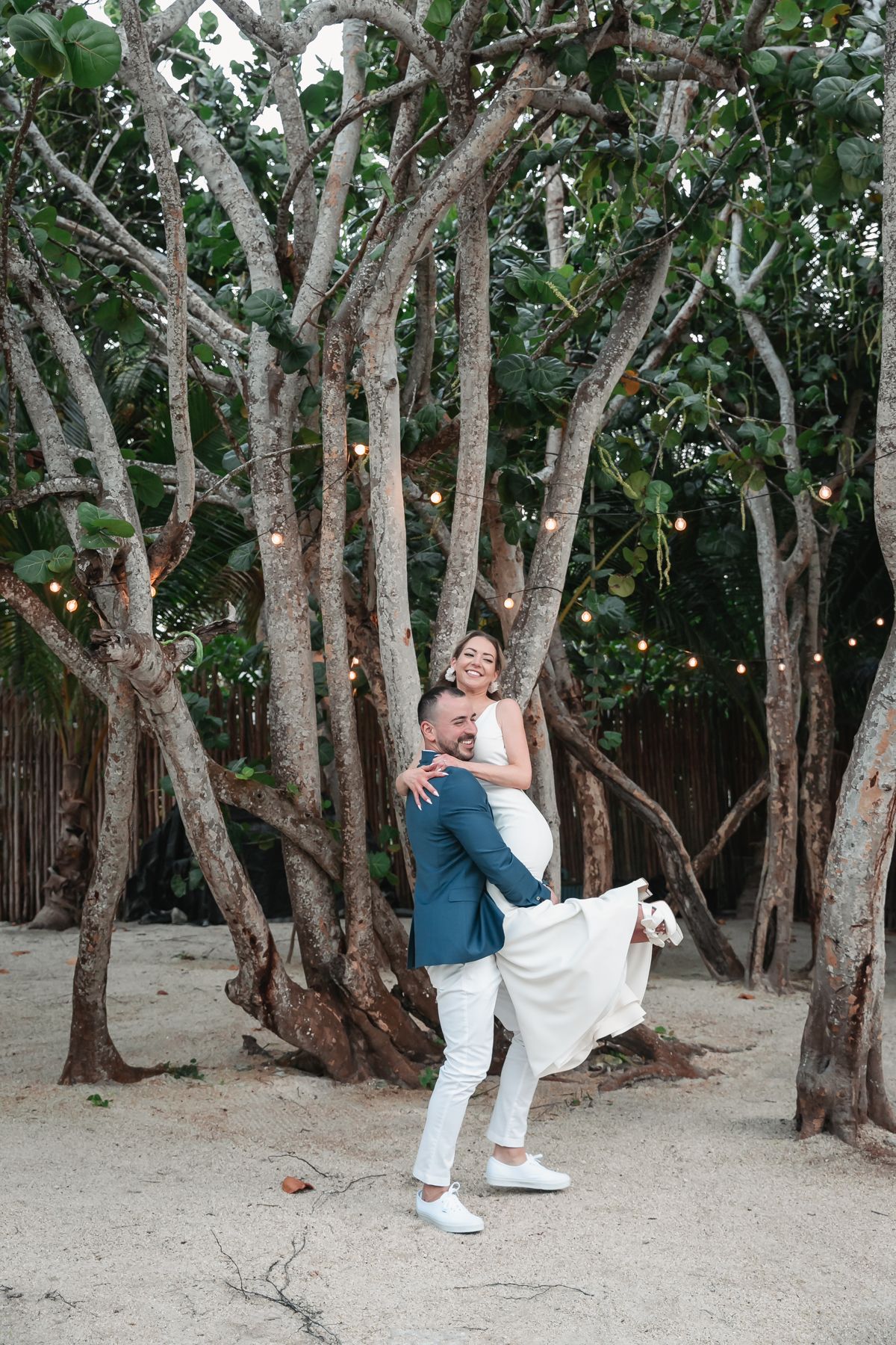Groom lifting smiling bride in white dress among tropical trees with string lights at sunset.