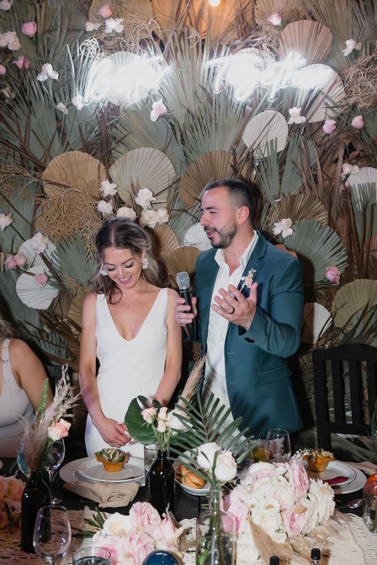 Bride and groom give a speech at their tropical wedding reception with a bohemian floral backdrop.