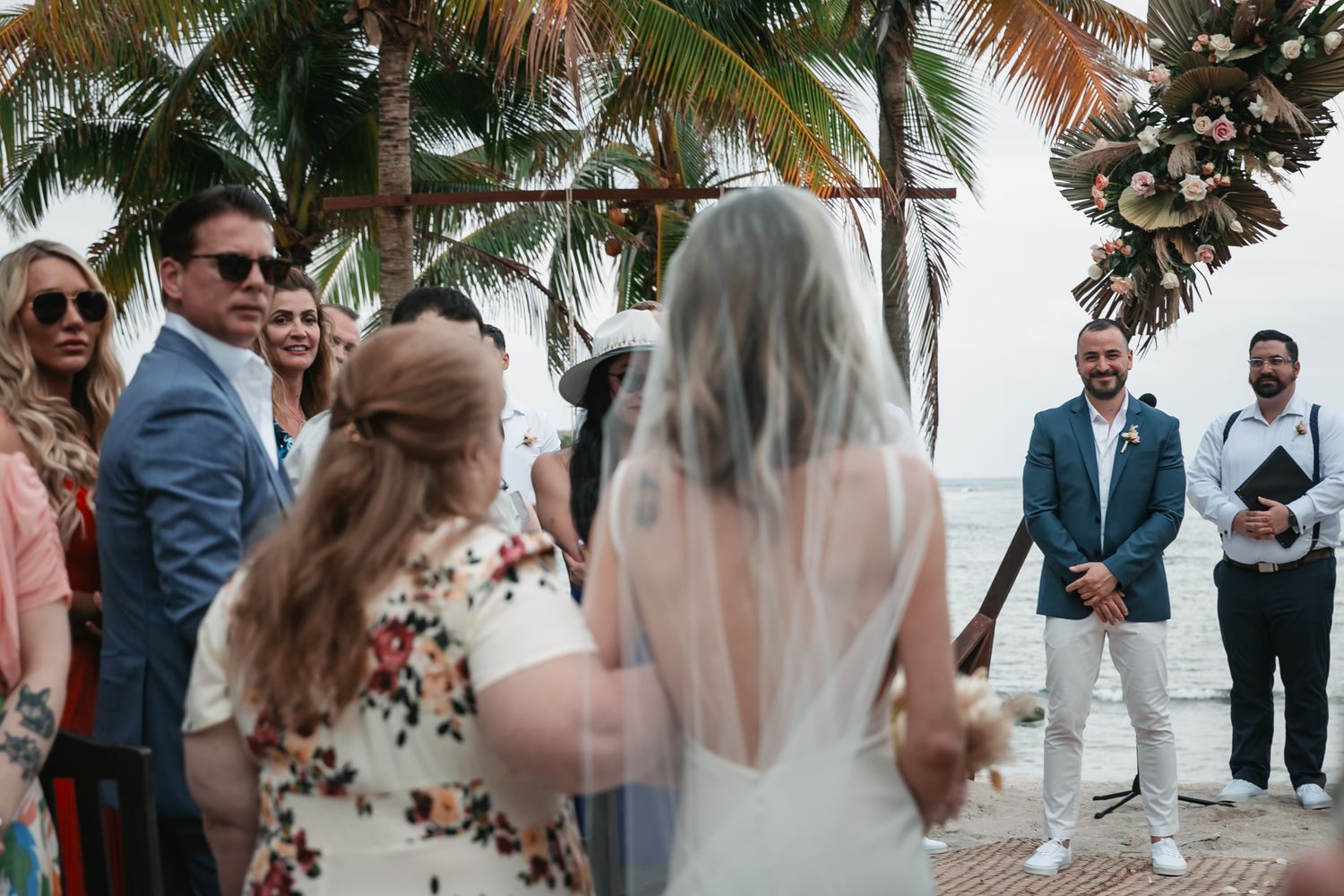 Bride in white gown and veil walks toward smiling groom at tropical beach wedding ceremony with guests watching.