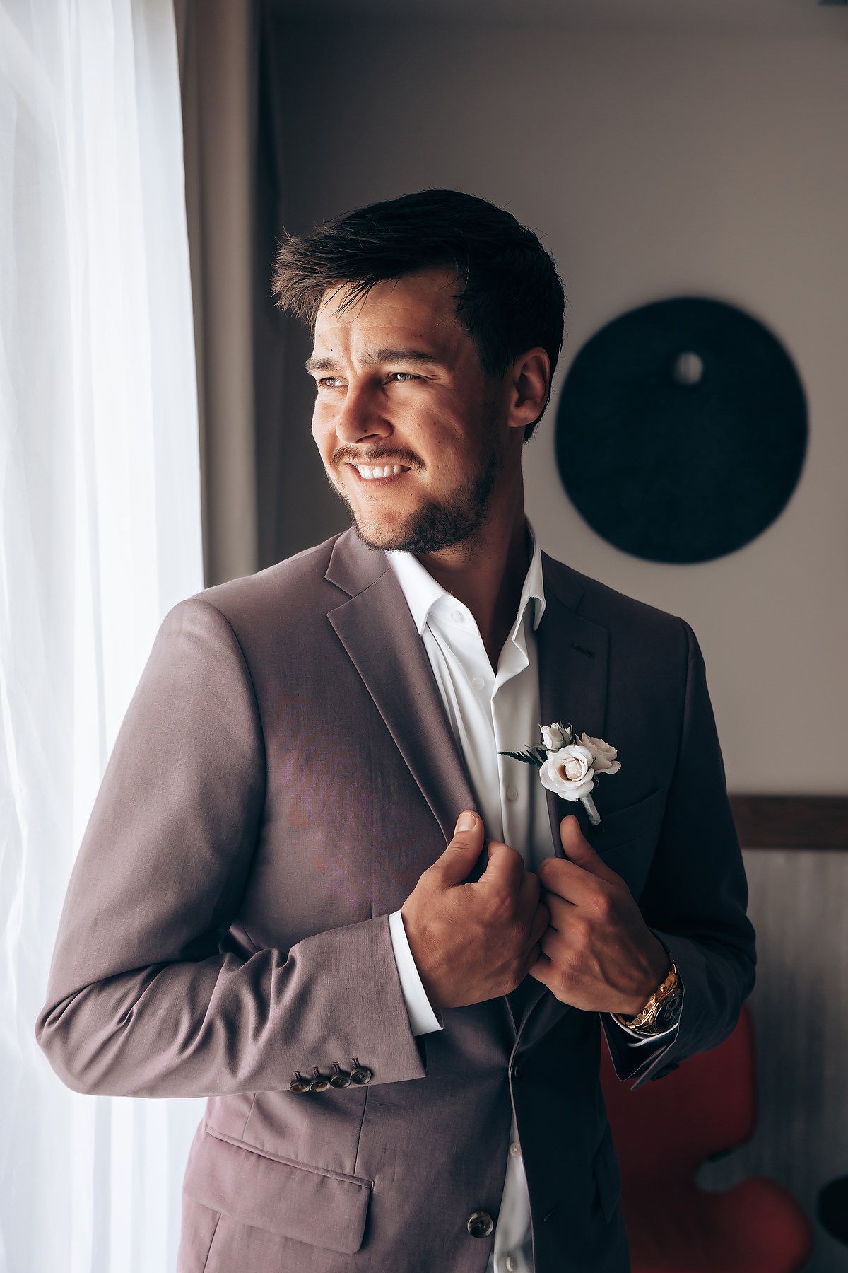 Groom in a light brown suit adjusts his boutonniere while standing near a window.