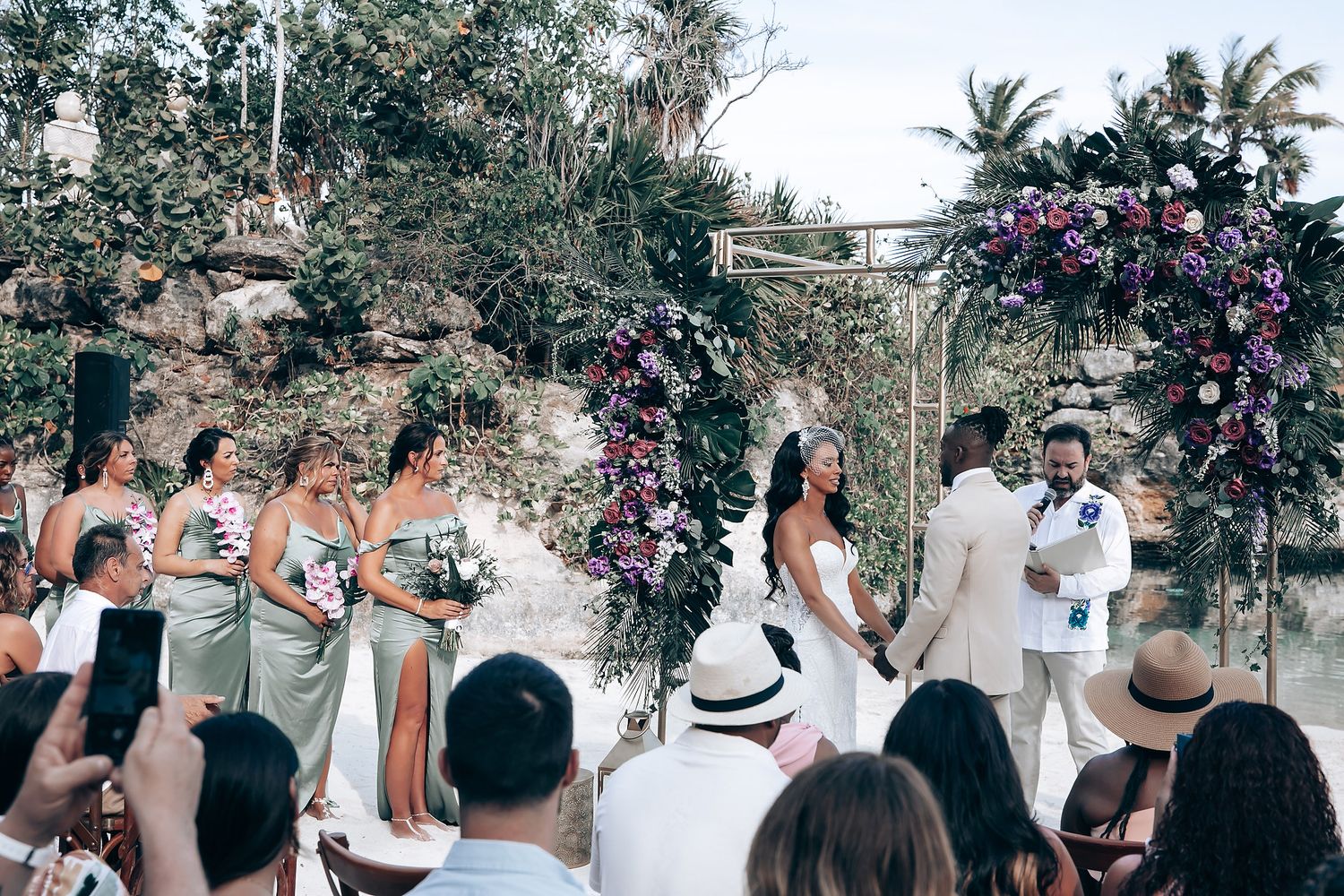 Outdoor wedding ceremony with guests watching couple exchange vows under floral-decorated arch.