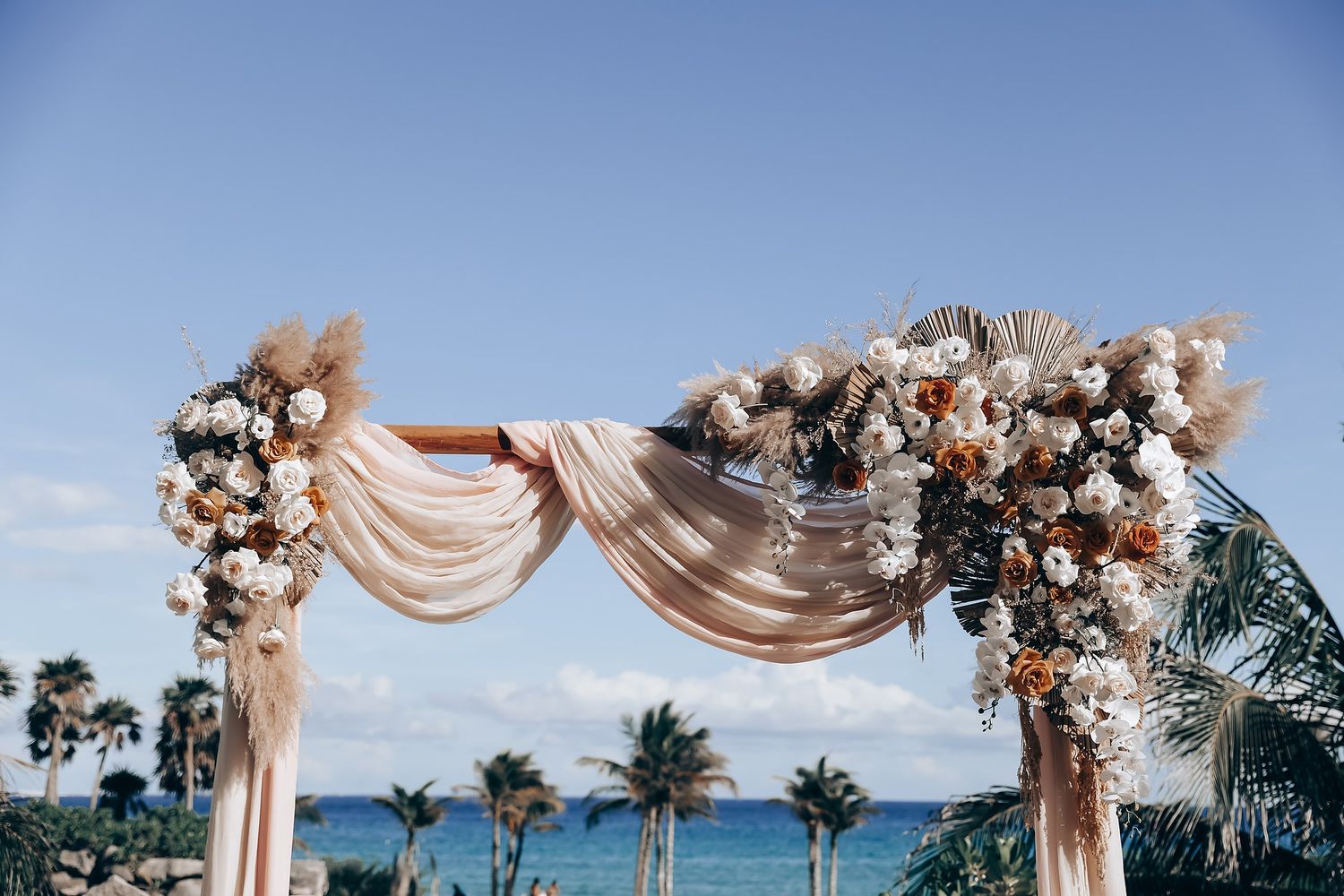 Elegant beach wedding ceremony setup with white draped fabric arch and floral arrangements overlooking tropical ocean views.