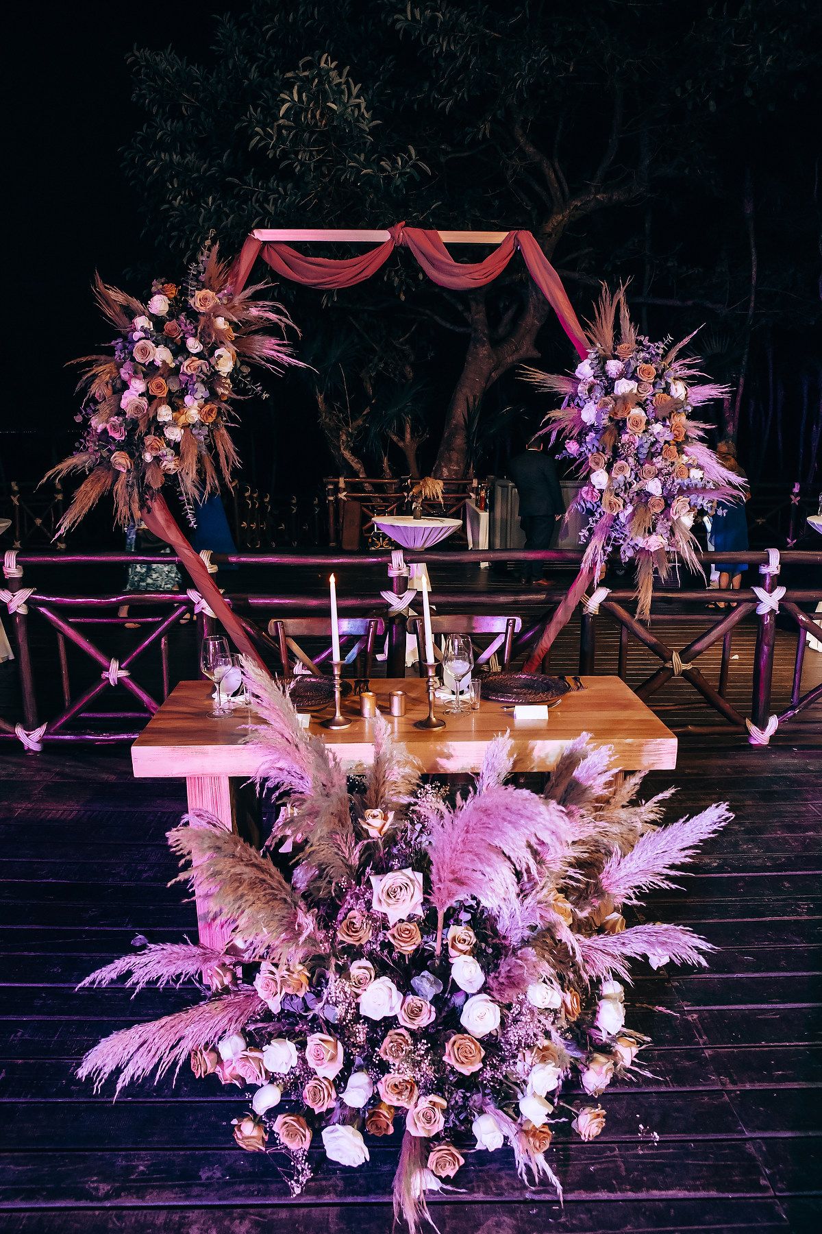 Elegant wedding decor featuring pink and purple floral arrangements with pampas grass and hanging lights in a dark venue.