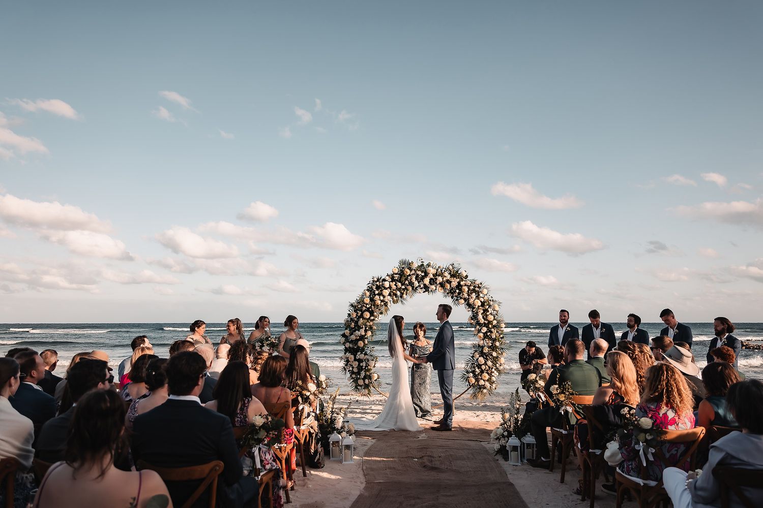 Beachside wedding ceremony with guests seated under floral arch and waves crashing in background.