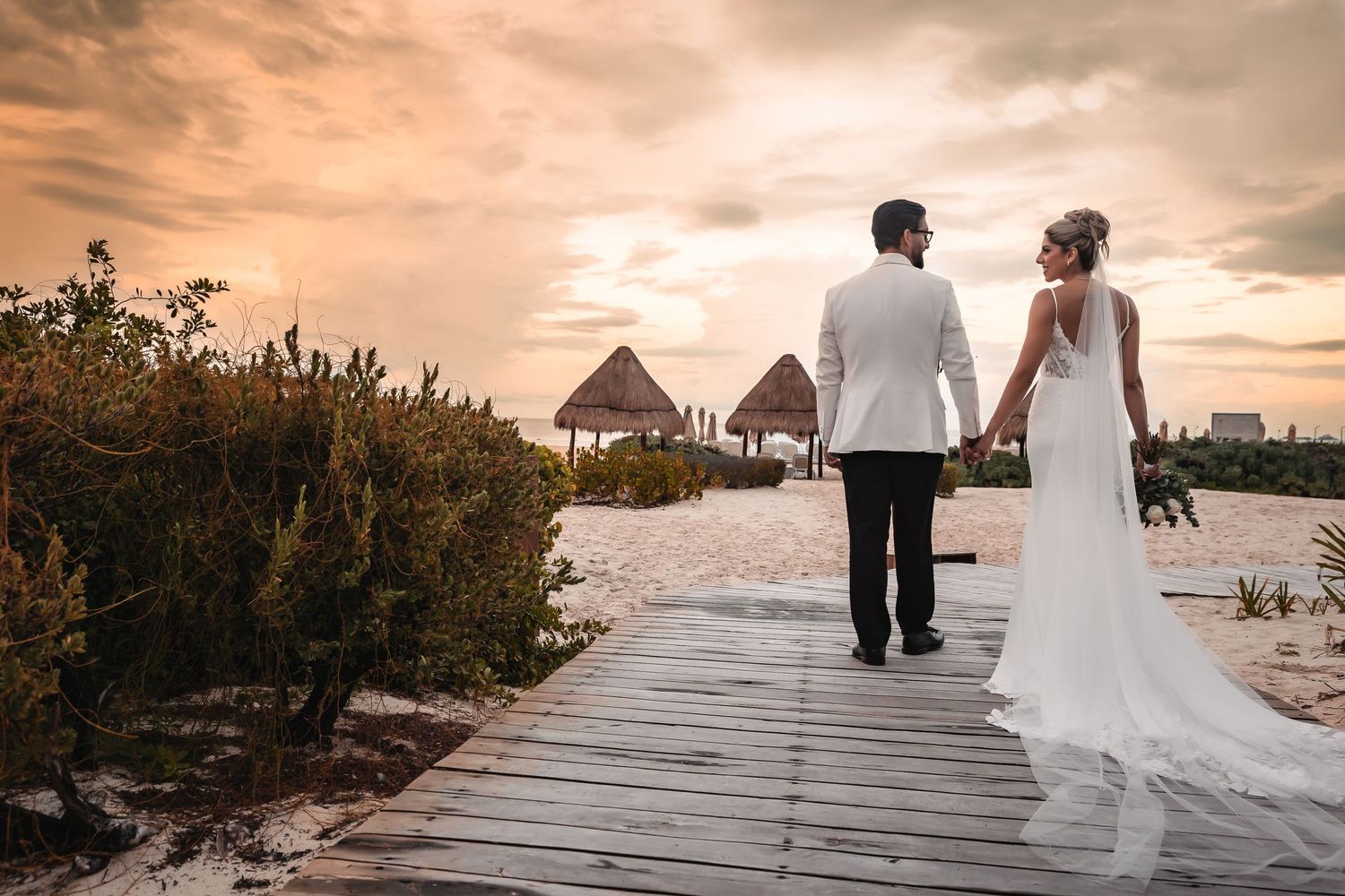 Romantic sunset wedding photo on wooden boardwalk with beach palapas in background.