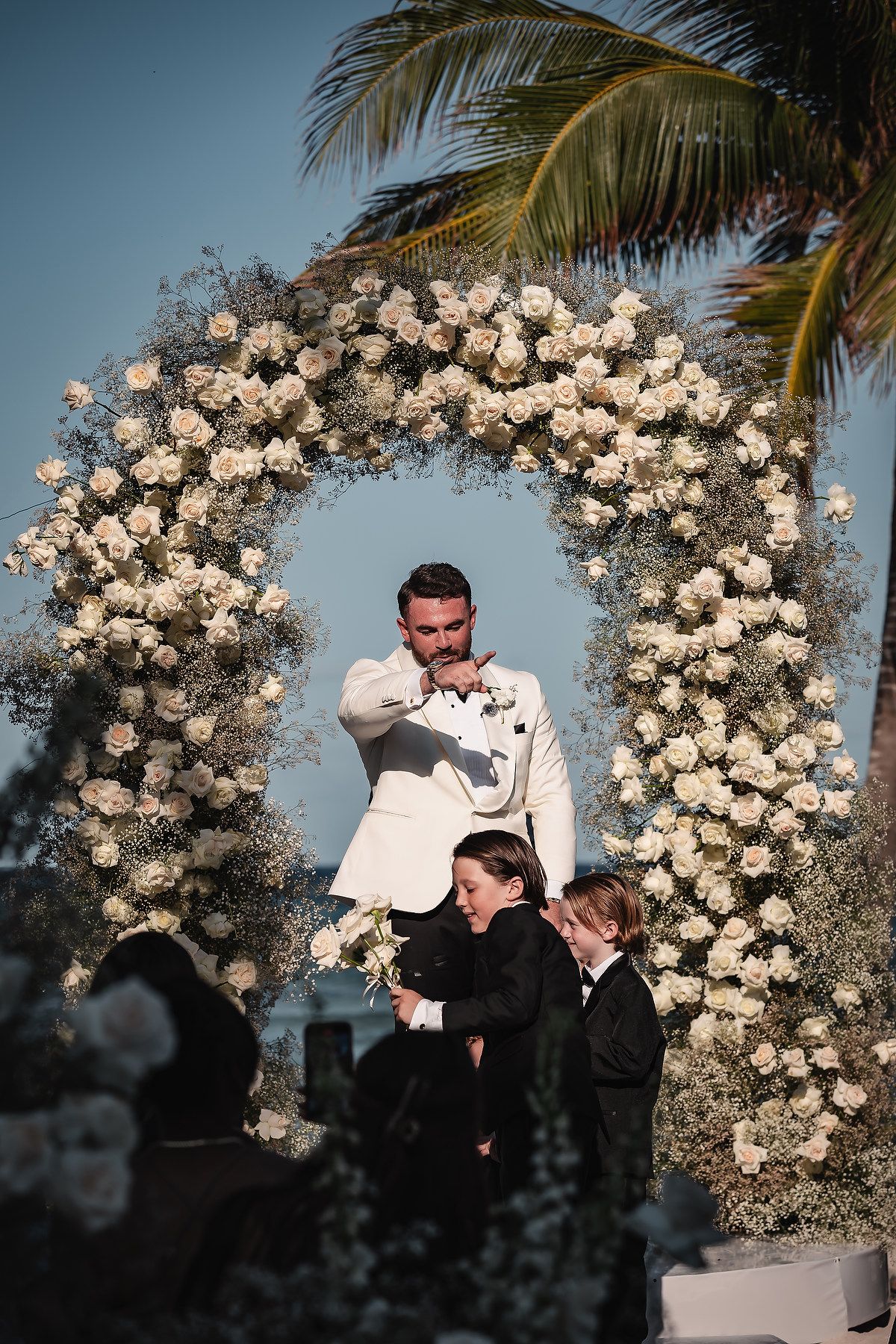 White floral arch frames an intimate moment during a beach wedding ceremony.