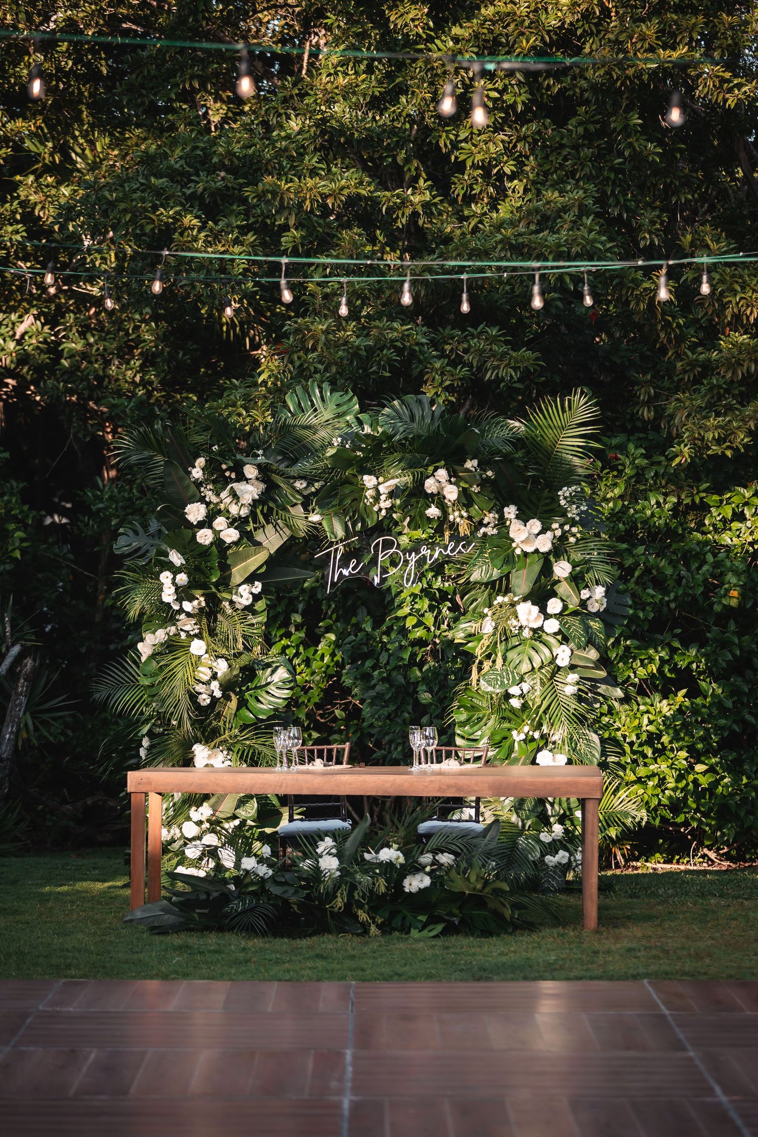 Outdoor wedding reception setup with string lights and floral arrangements on wooden tables.