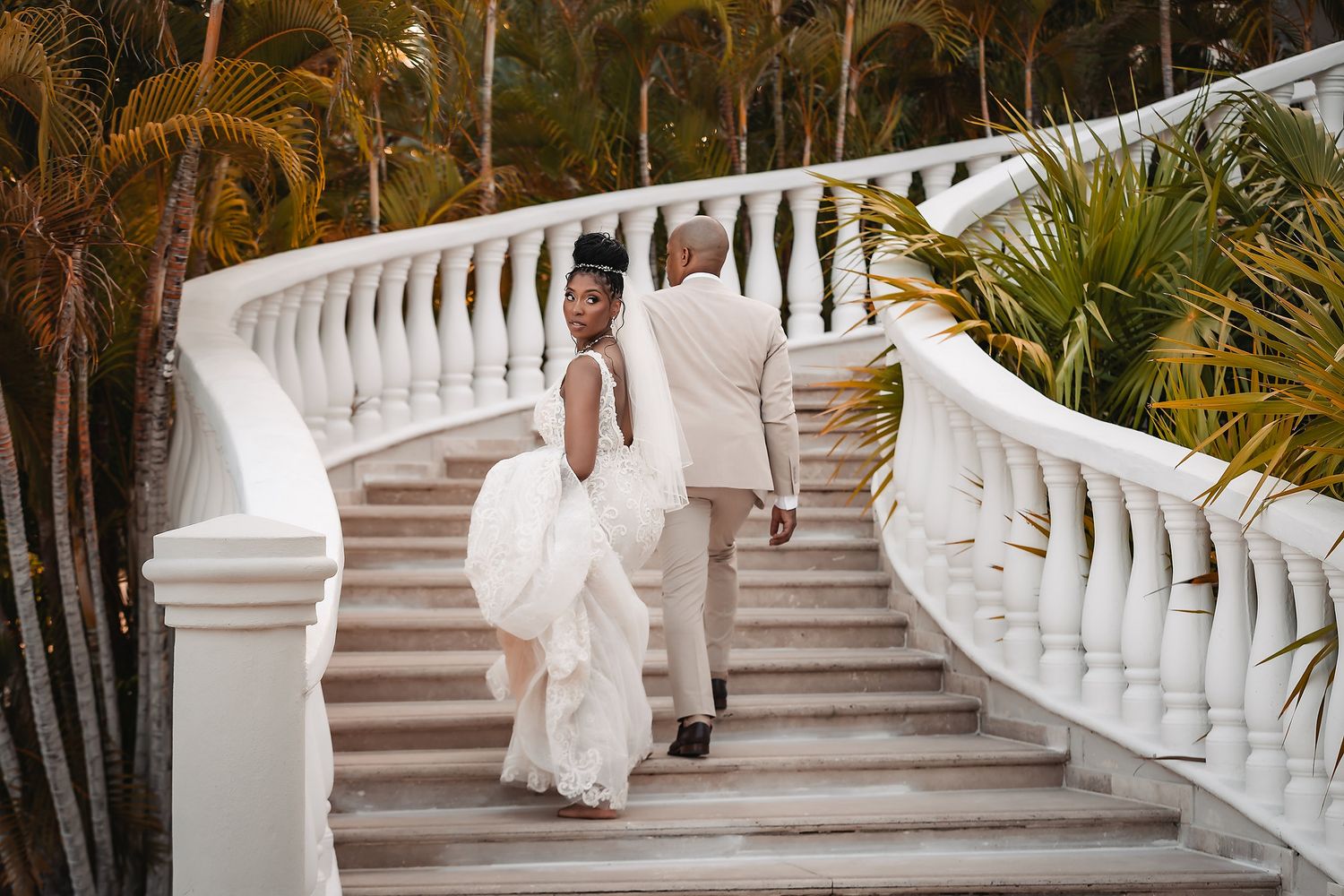 A bride descends elegant white stone stairs surrounded by tropical landscaping.