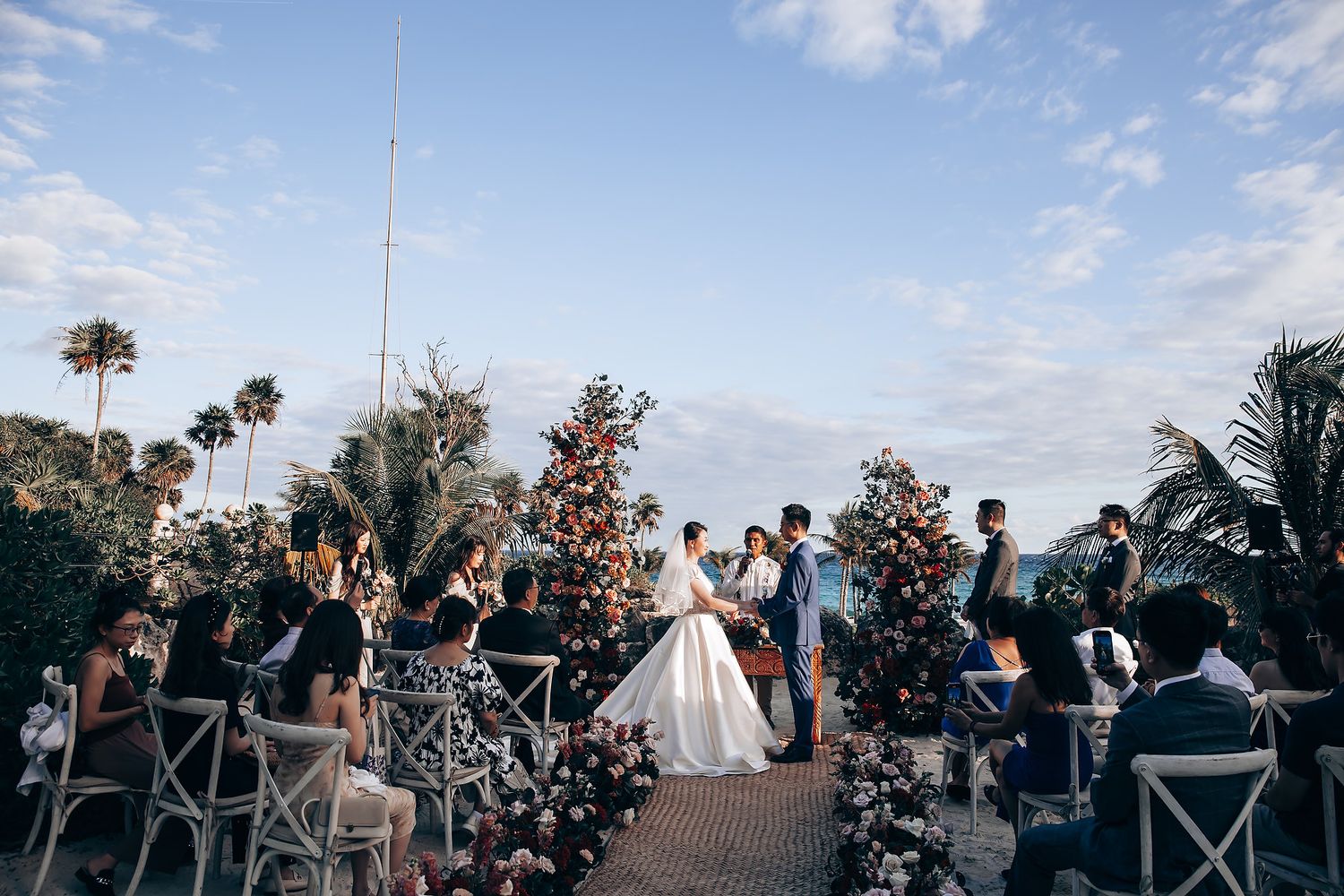 Outdoor wedding ceremony setup with floral arrangements and cross-back chairs under blue sky.