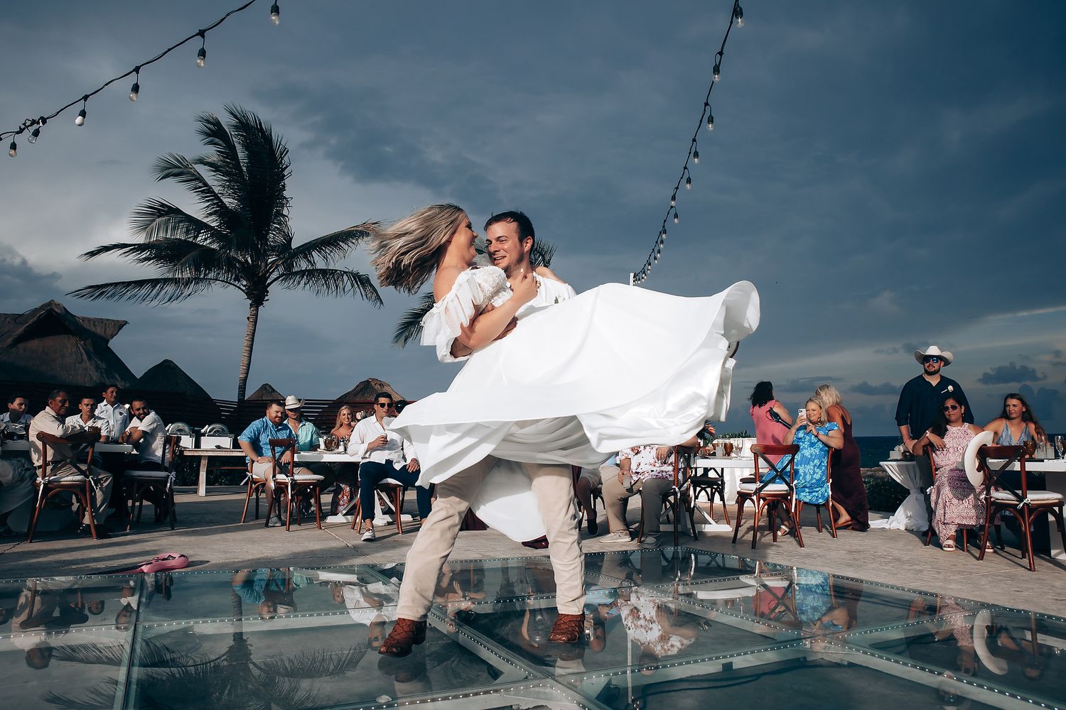 Wedding reception dance floor by the ocean with string lights and palm trees at dusk.
