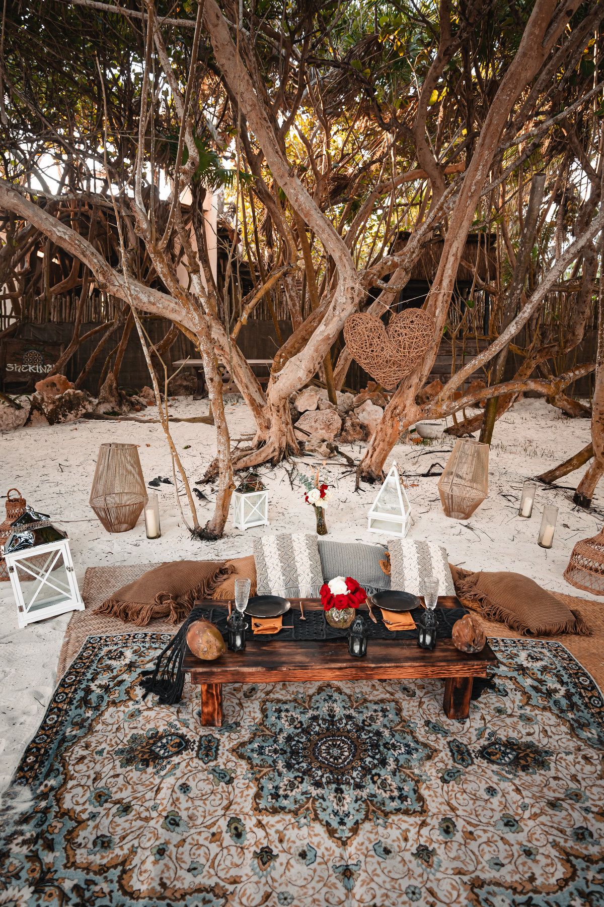 Intimate beach dining setup with rustic wooden table and decorative lanterns under trees.