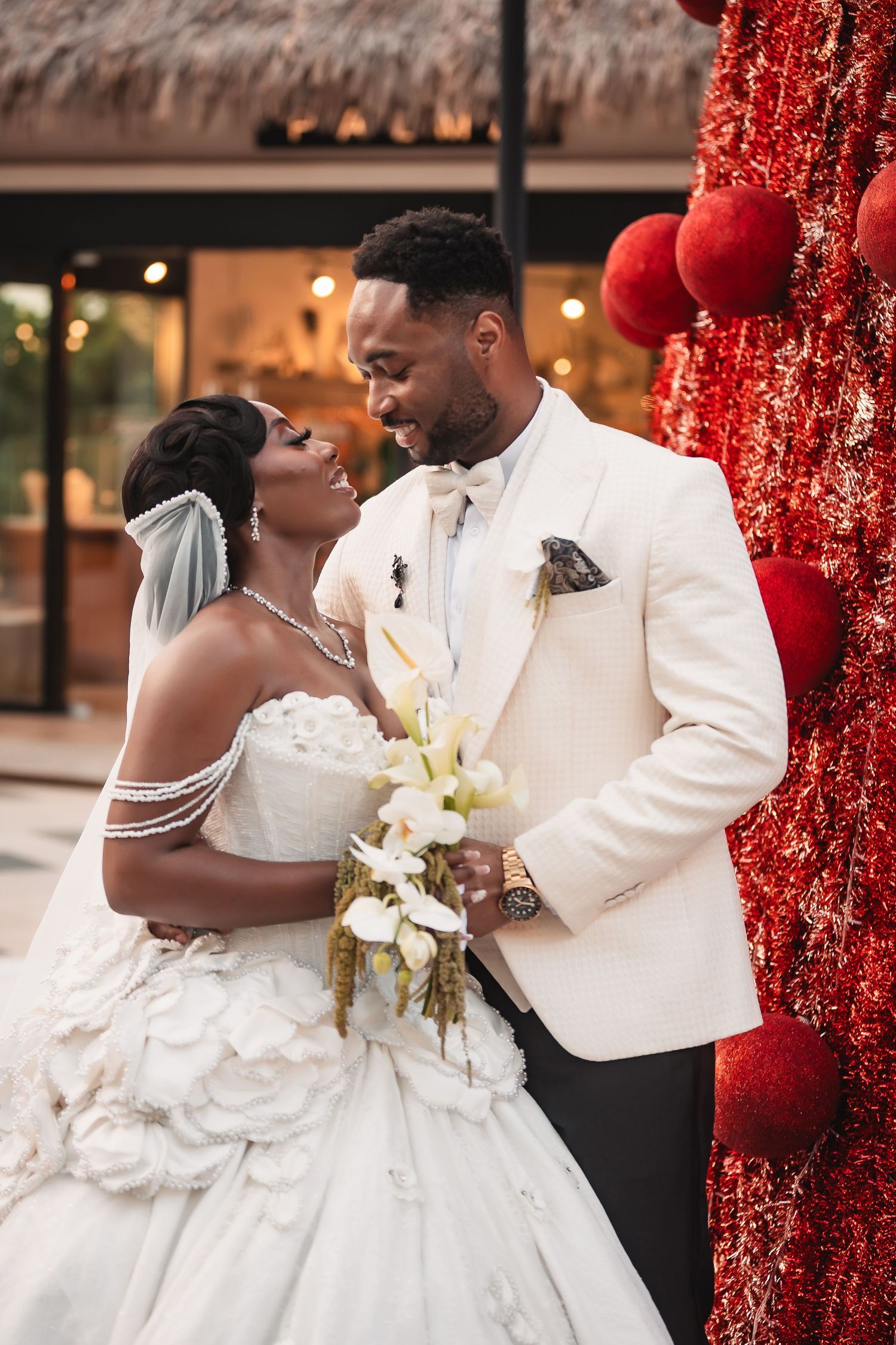 Elegant wedding couple poses against a festive red backdrop with white floral bouquet.