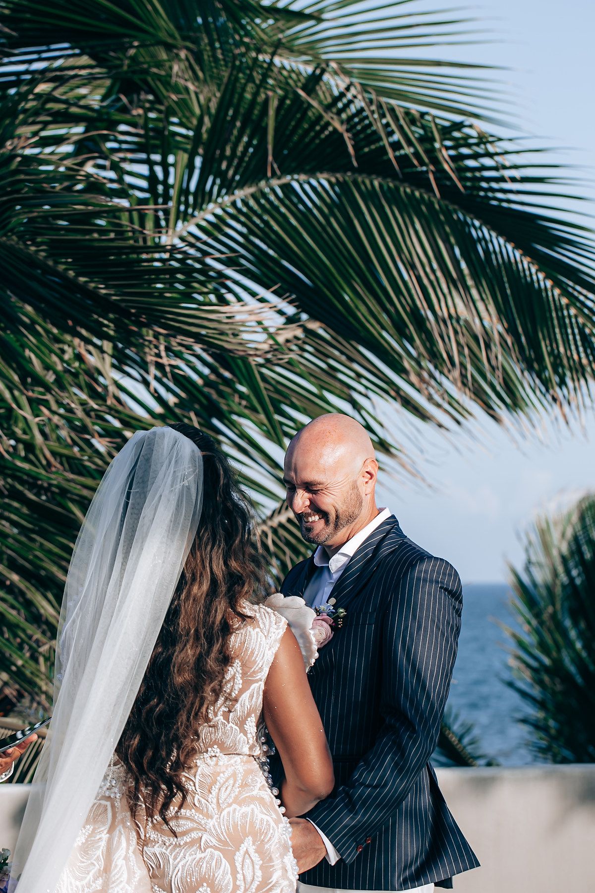 A couple shares a romantic moment under swaying palm fronds at a tropical beach wedding ceremony.