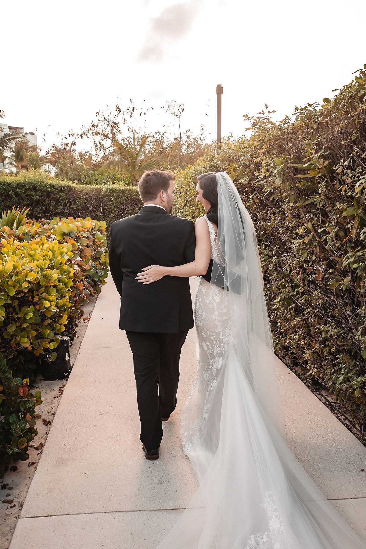 Couple strolls hand in hand down a sunlit pathway surrounded by tropical landscaping.