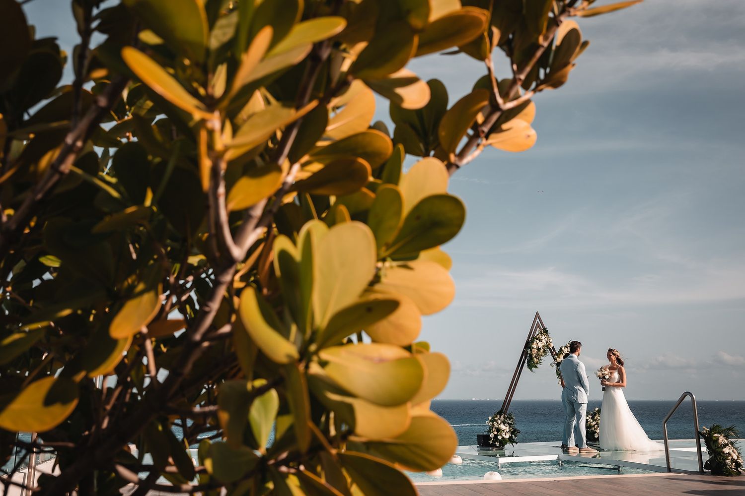 Tropical foliage frames an oceanside wedding ceremony with geometric arch and white chairs.
