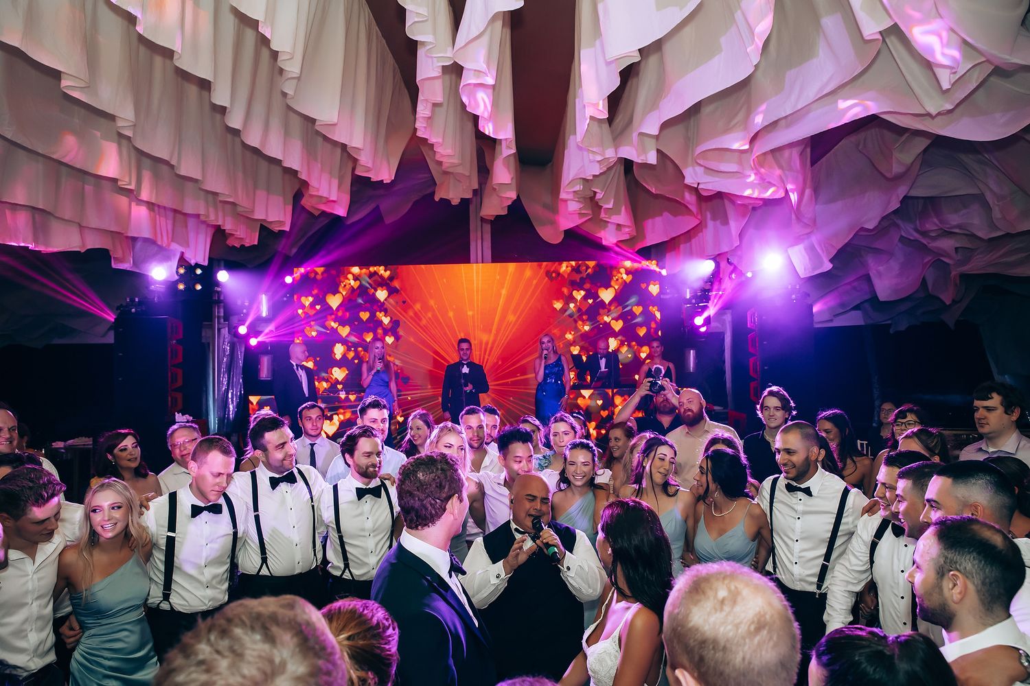 Wedding guests dancing and celebrating on a colorful dance floor illuminated by vibrant purple and orange lighting.